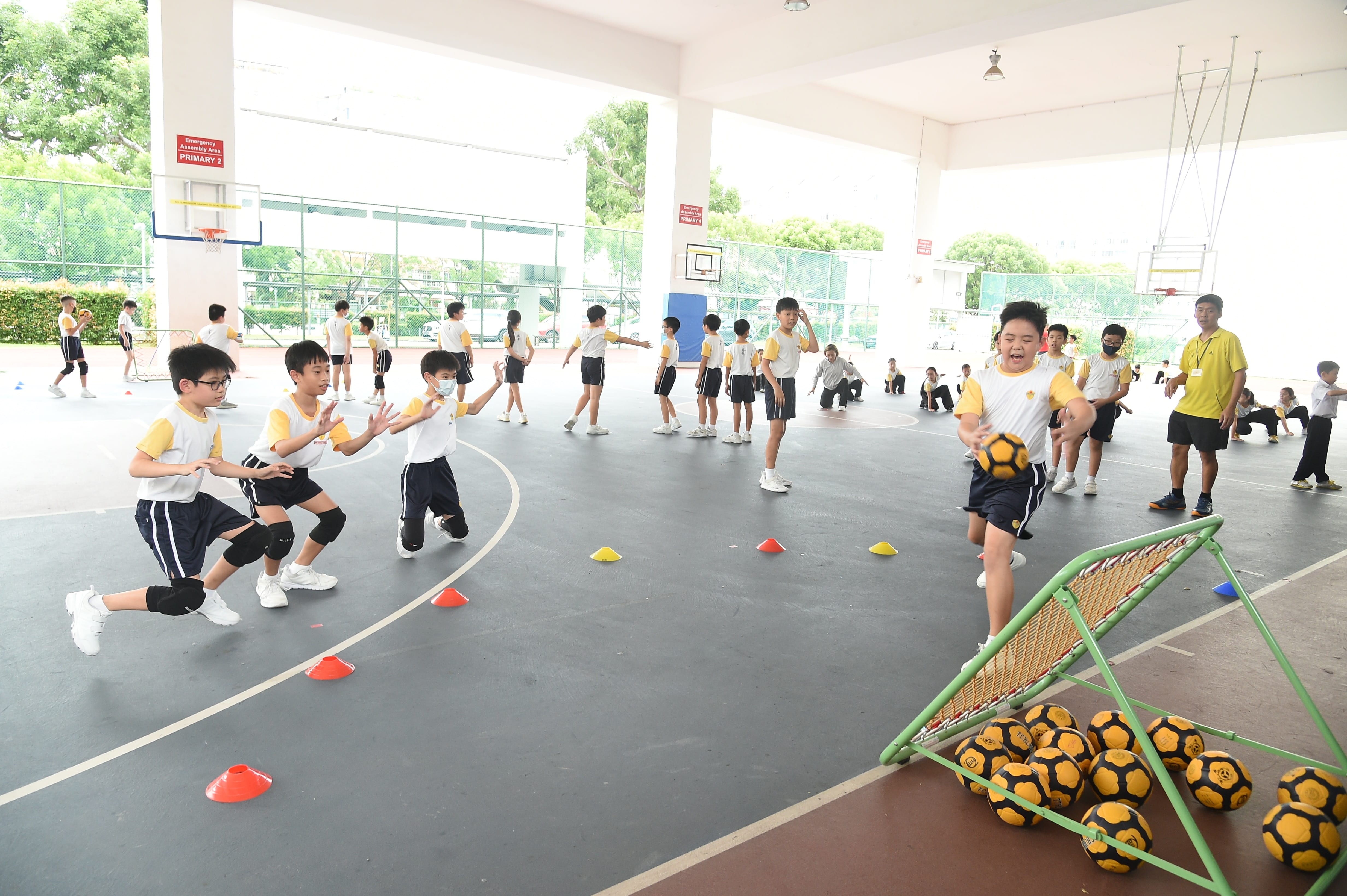 Children in white and yellow uniforms doing Tchoukball drills on a covered school court with footballs and cones.