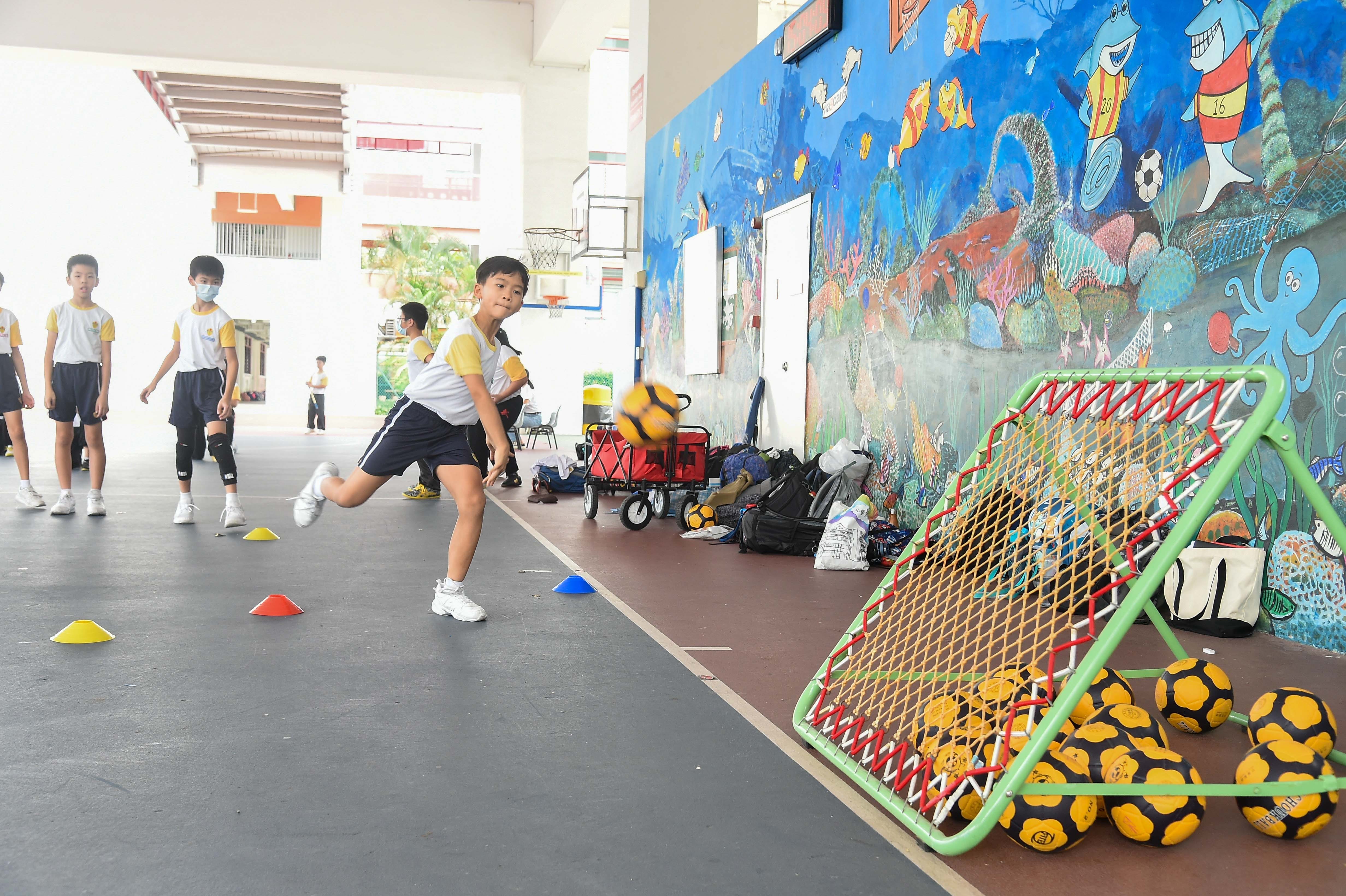 A child throws a ball toward a net during a school PE class, with colorful ocean mural and other students in the background.