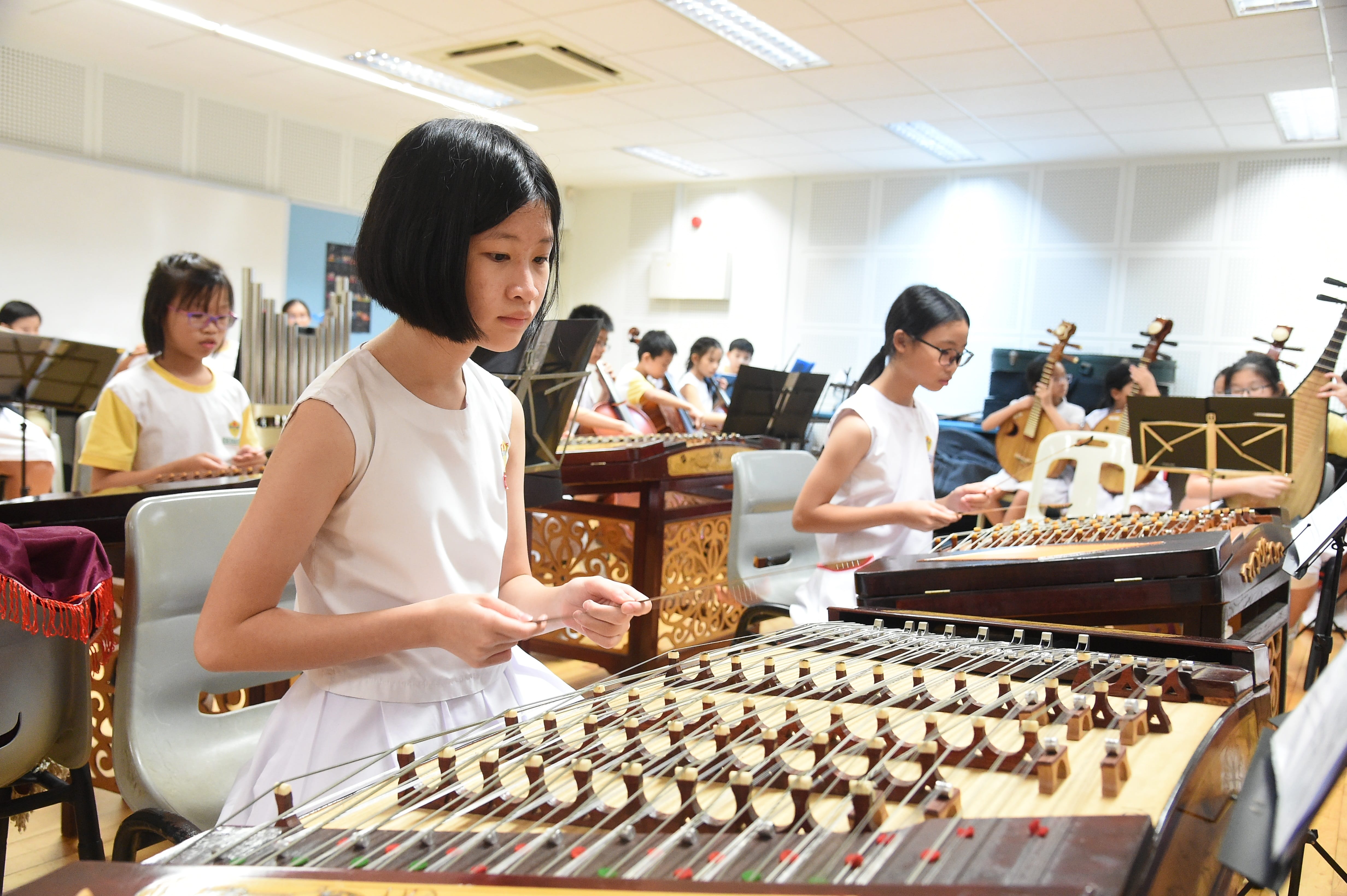 Several children in white uniforms play traditional Chinese string instruments in a bright music classroom.