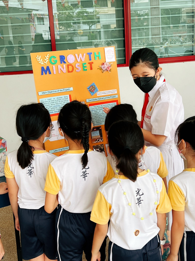 A student presents a colorful Growth Mindset poster to a group of younger uniformed schoolchildren.