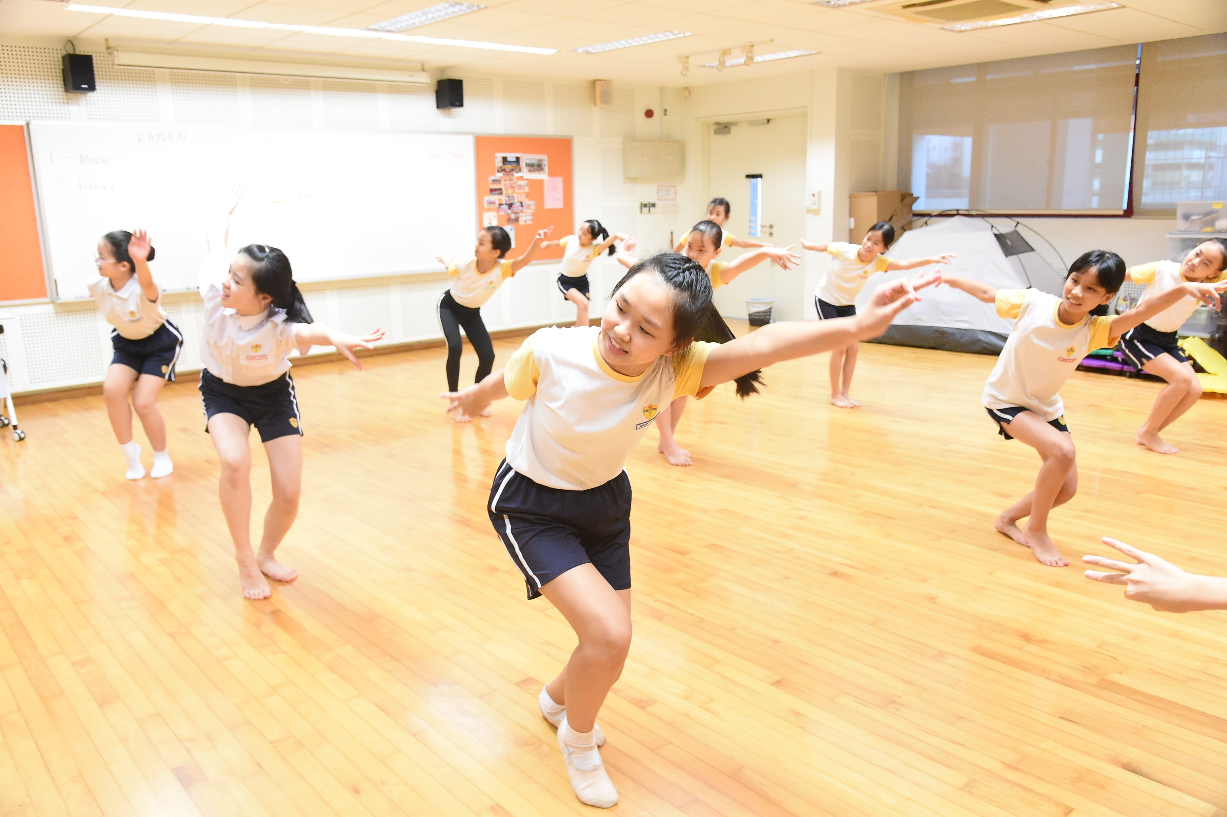 A group of young girls in school uniforms practice a dance routine on a wooden floor in a bright indoor room.