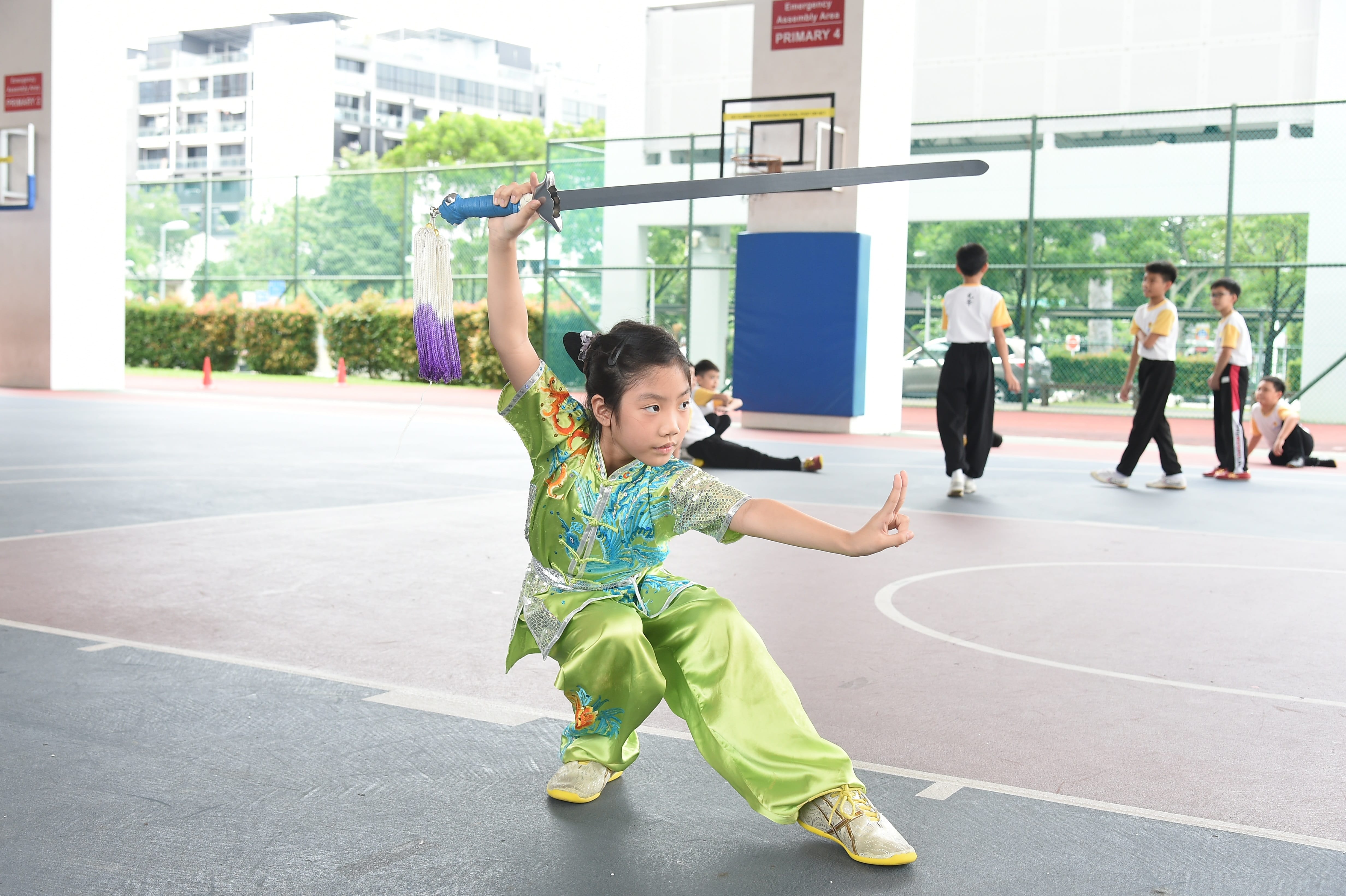 A girl in a green wushu uniform performs a sword pose on a school court with other children practicing in the background.