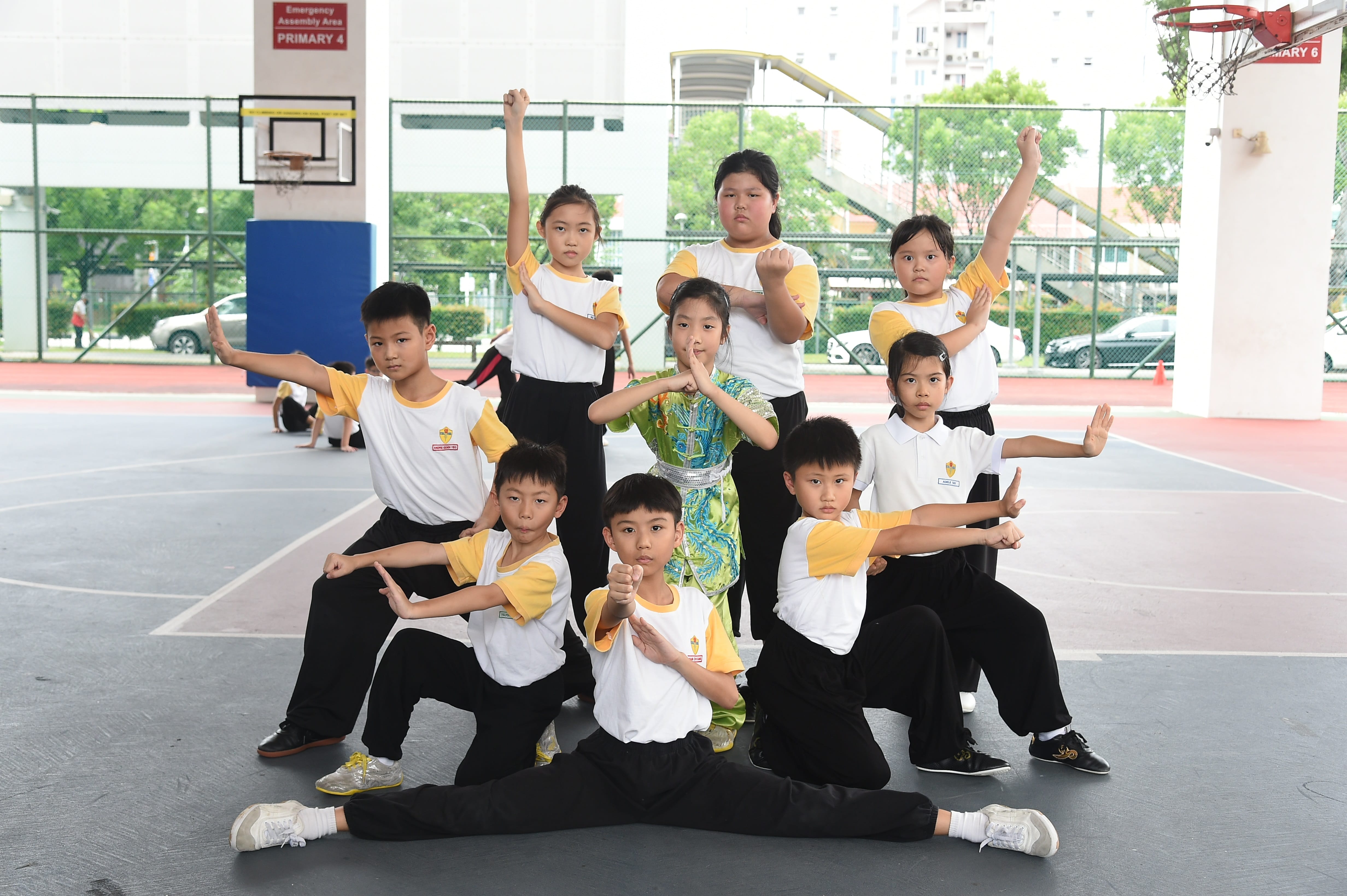 Nine children in white and yellow uniforms pose in martial arts stances on a school basketball court.