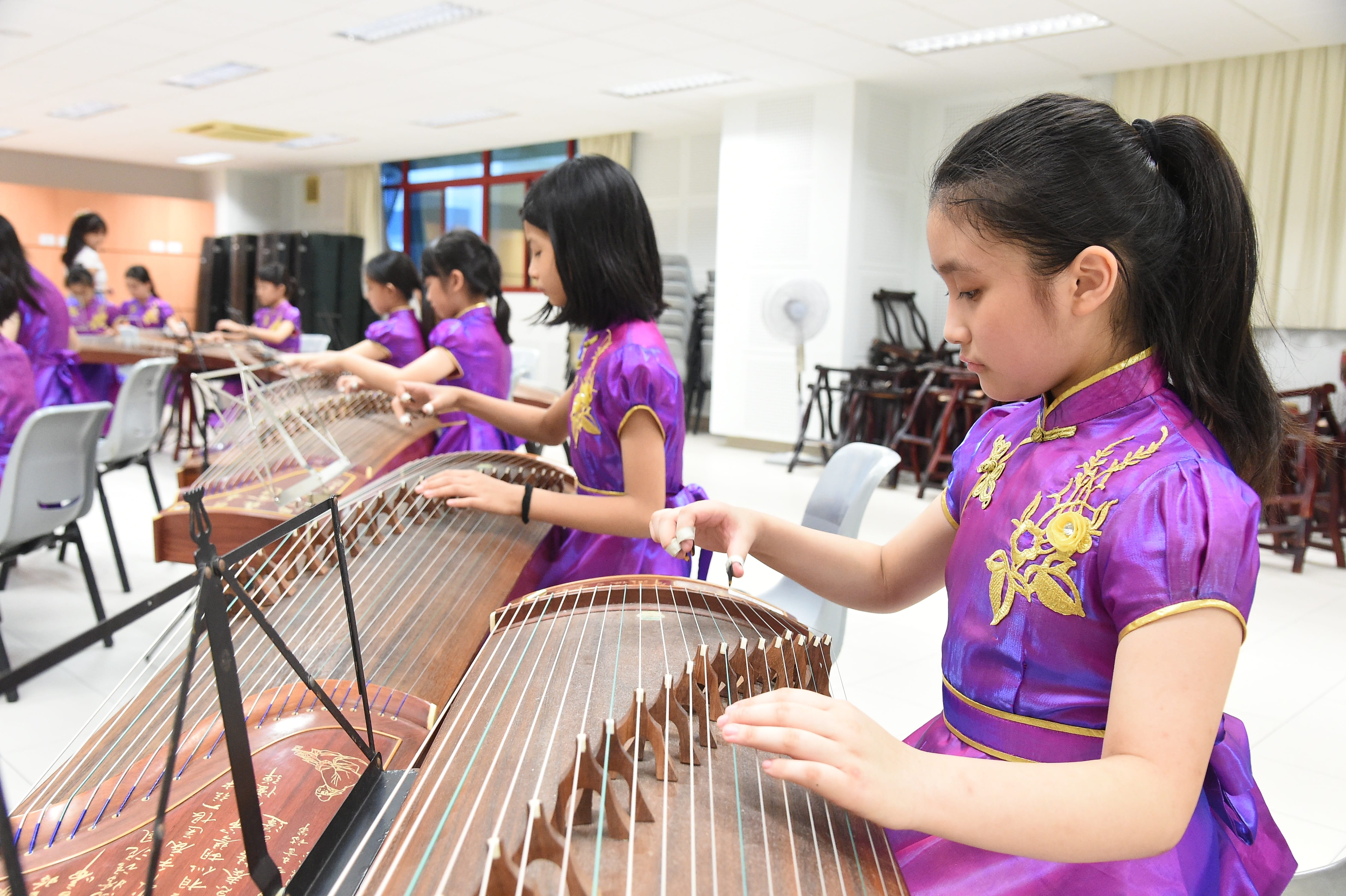 Several girls in purple embroidered dresses play guzheng instruments in a classroom setting.
