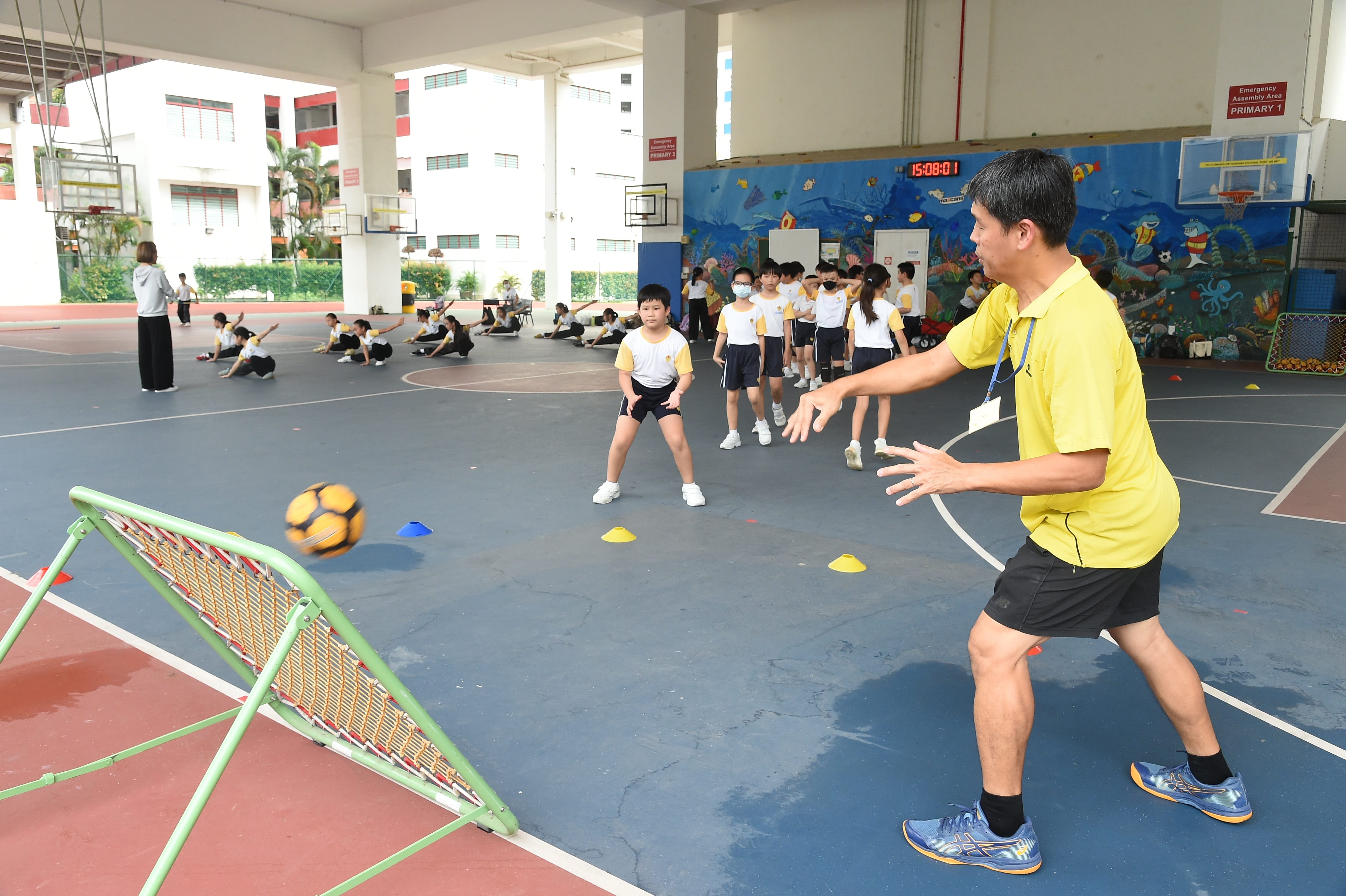 An instructor in yellow passes a ball to a child on a school court while other students watch or stretch nearby.