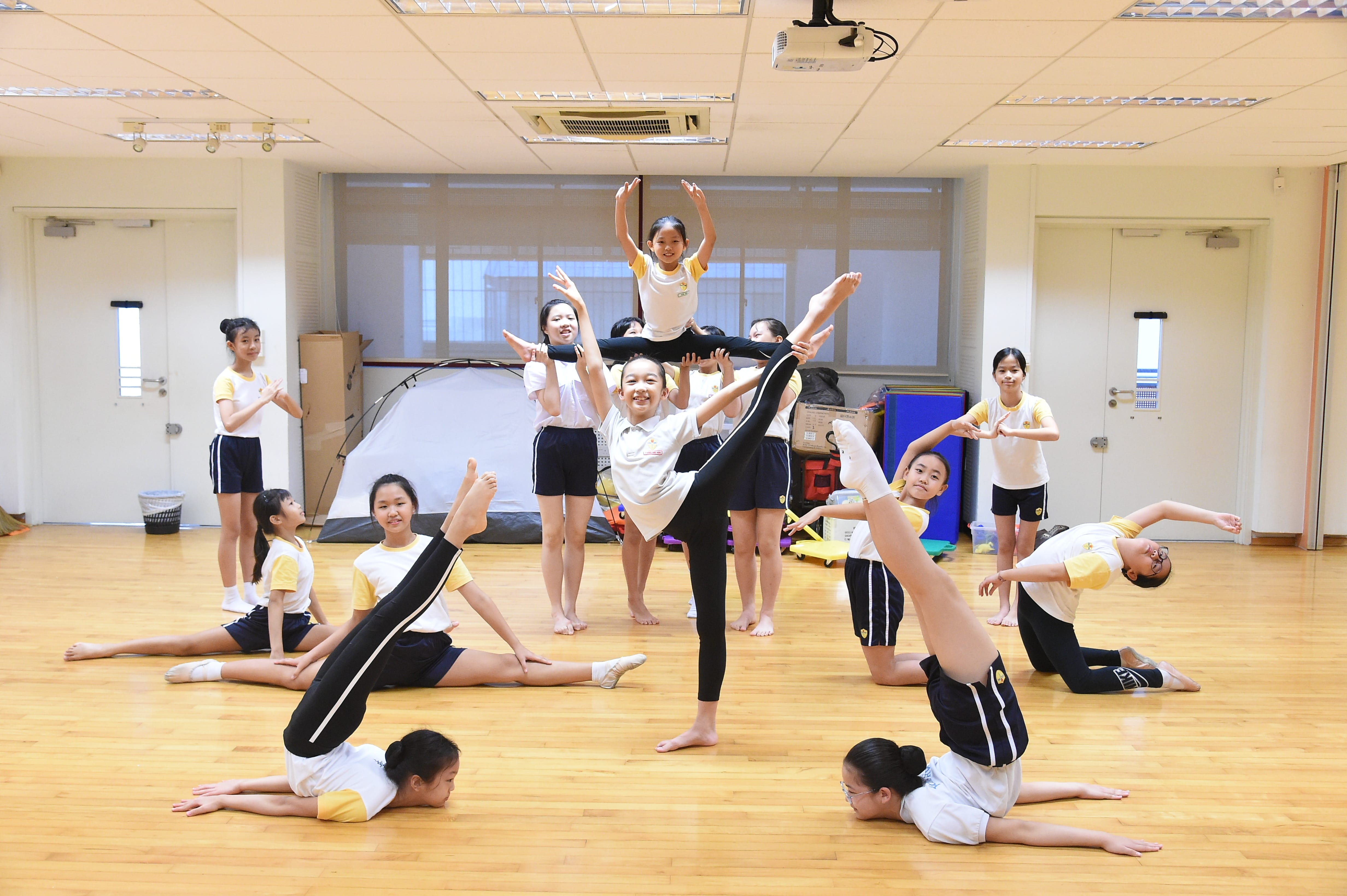 A group of young girls in uniforms performing acrobatic and gymnastics poses in an indoor studio with wood floors.