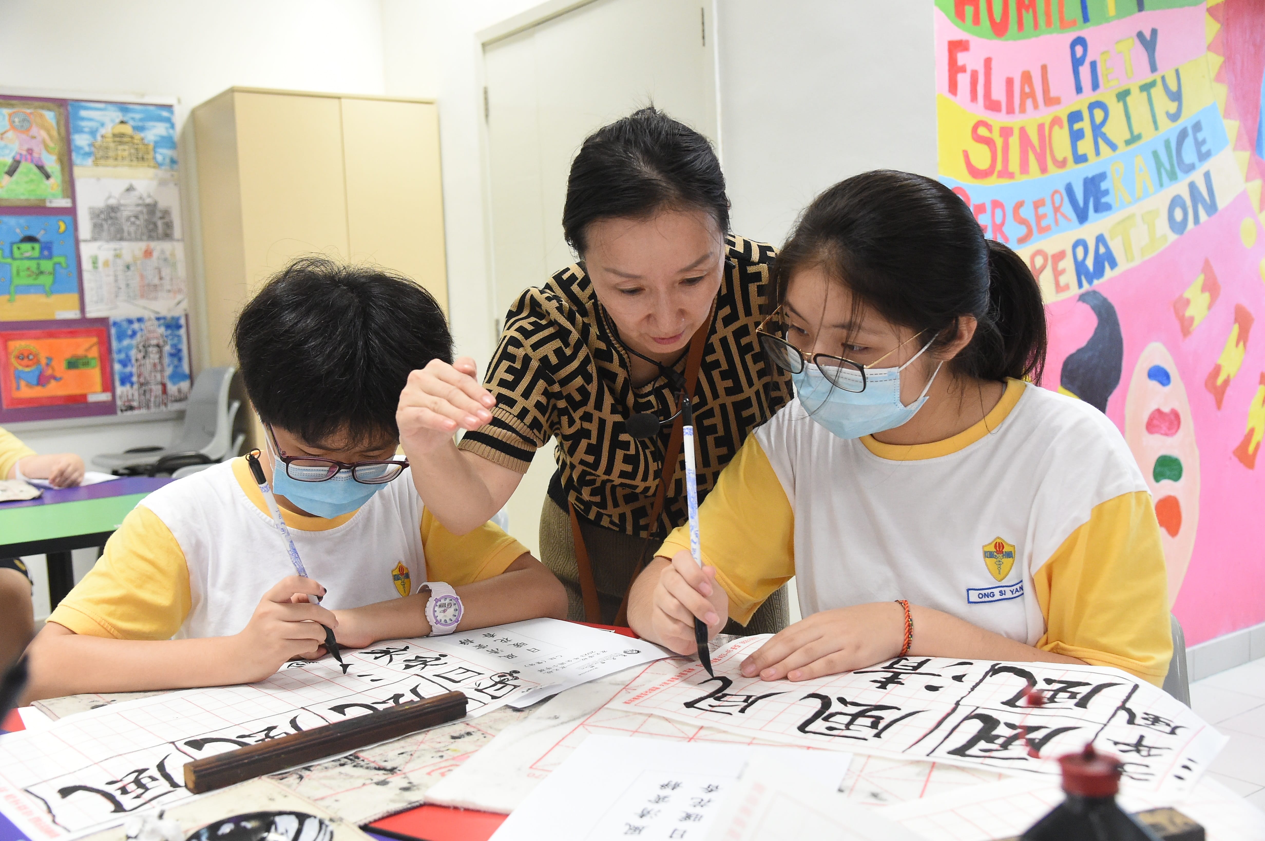 A teacher guides two masked students practicing Chinese calligraphy with ink brushes in a colorful classroom.