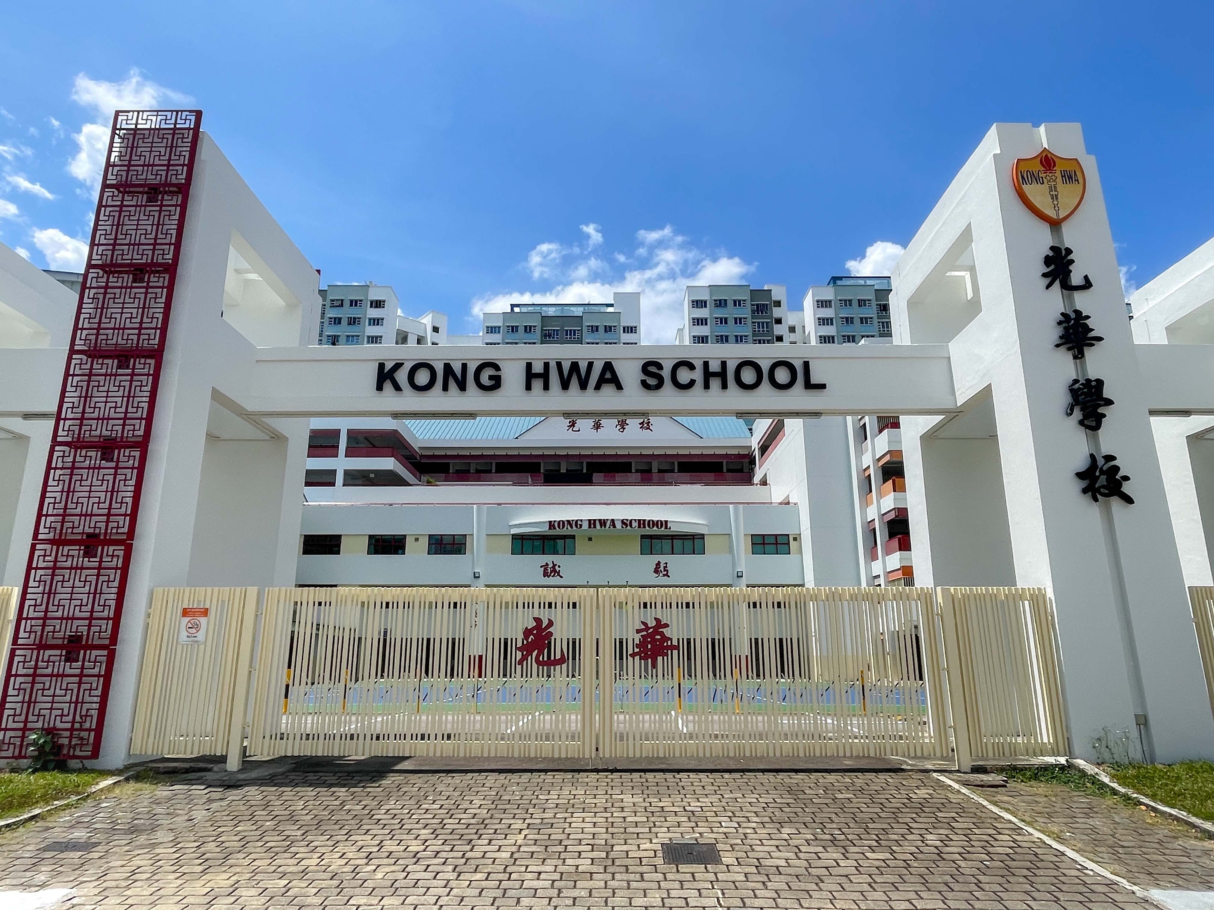 White entrance gate of Kong Hwa School with Chinese characters, closed sliding gates, and school buildings behind under blue sky.