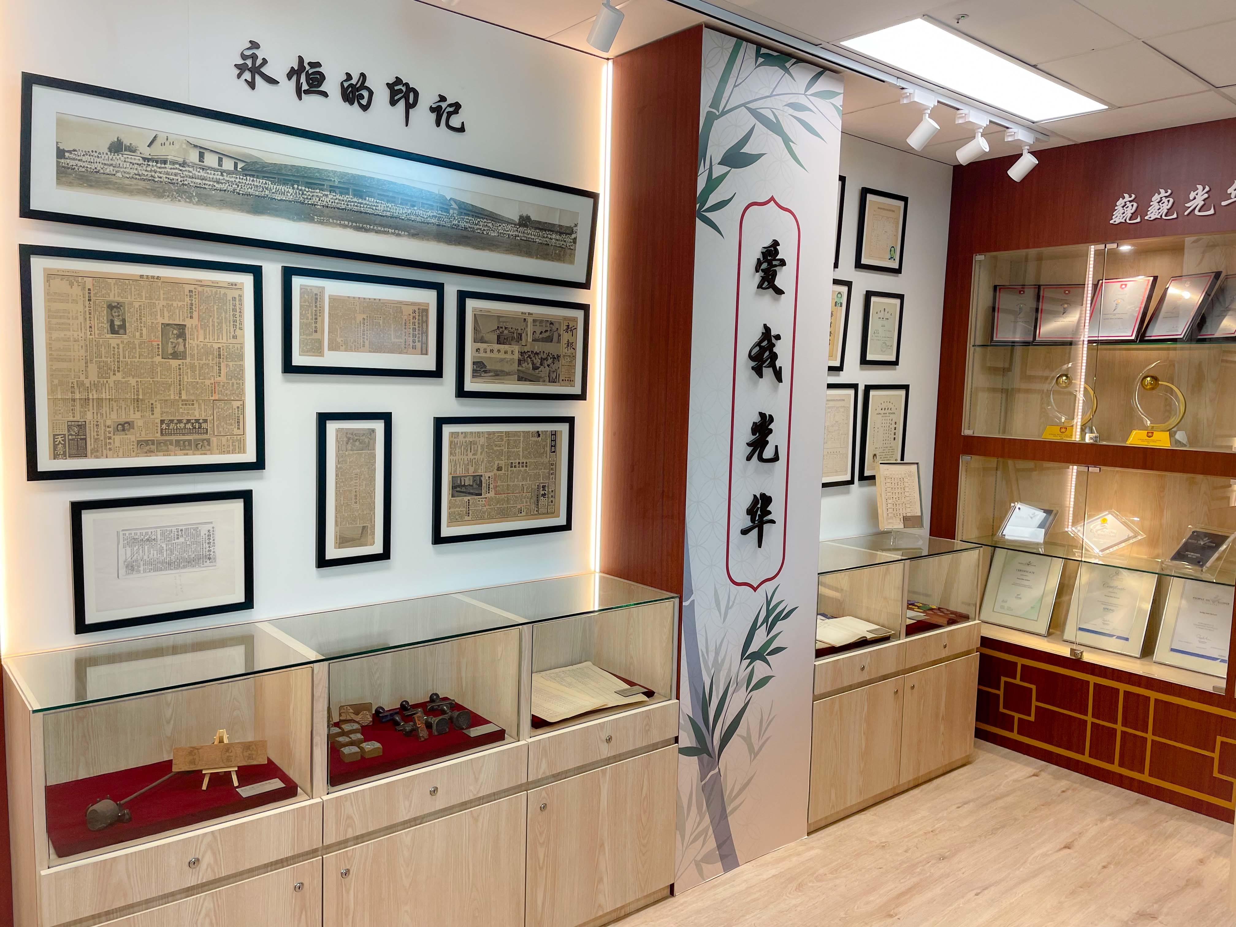 A Chinese museum display room with framed newspaper clippings, artifacts in glass cases, awards, and calligraphy signage on walls.