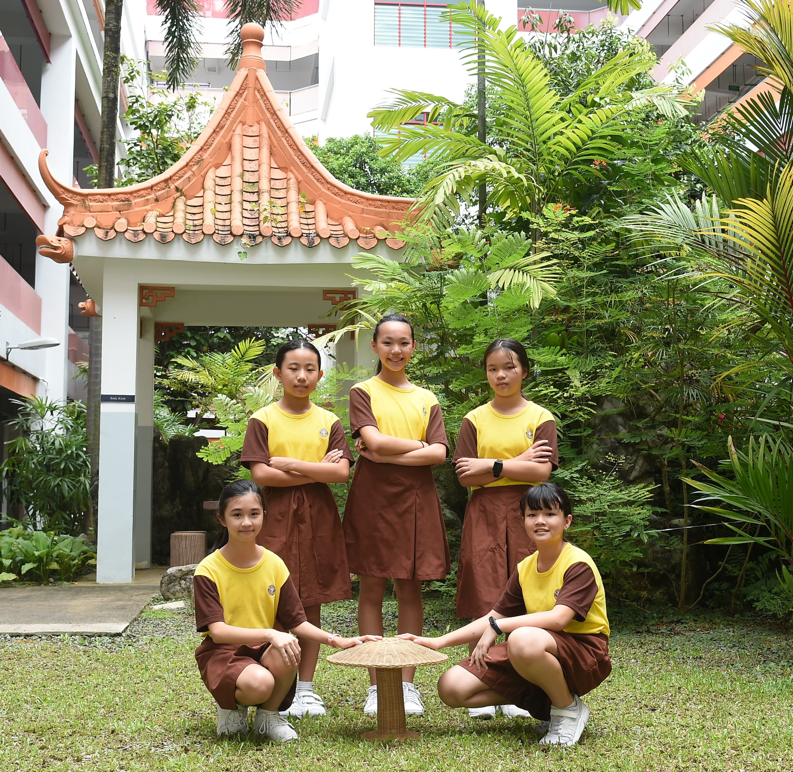 Five girls in yellow and brown school uniforms pose outdoors near a Chinese-style pavilion gate with lush greenery.