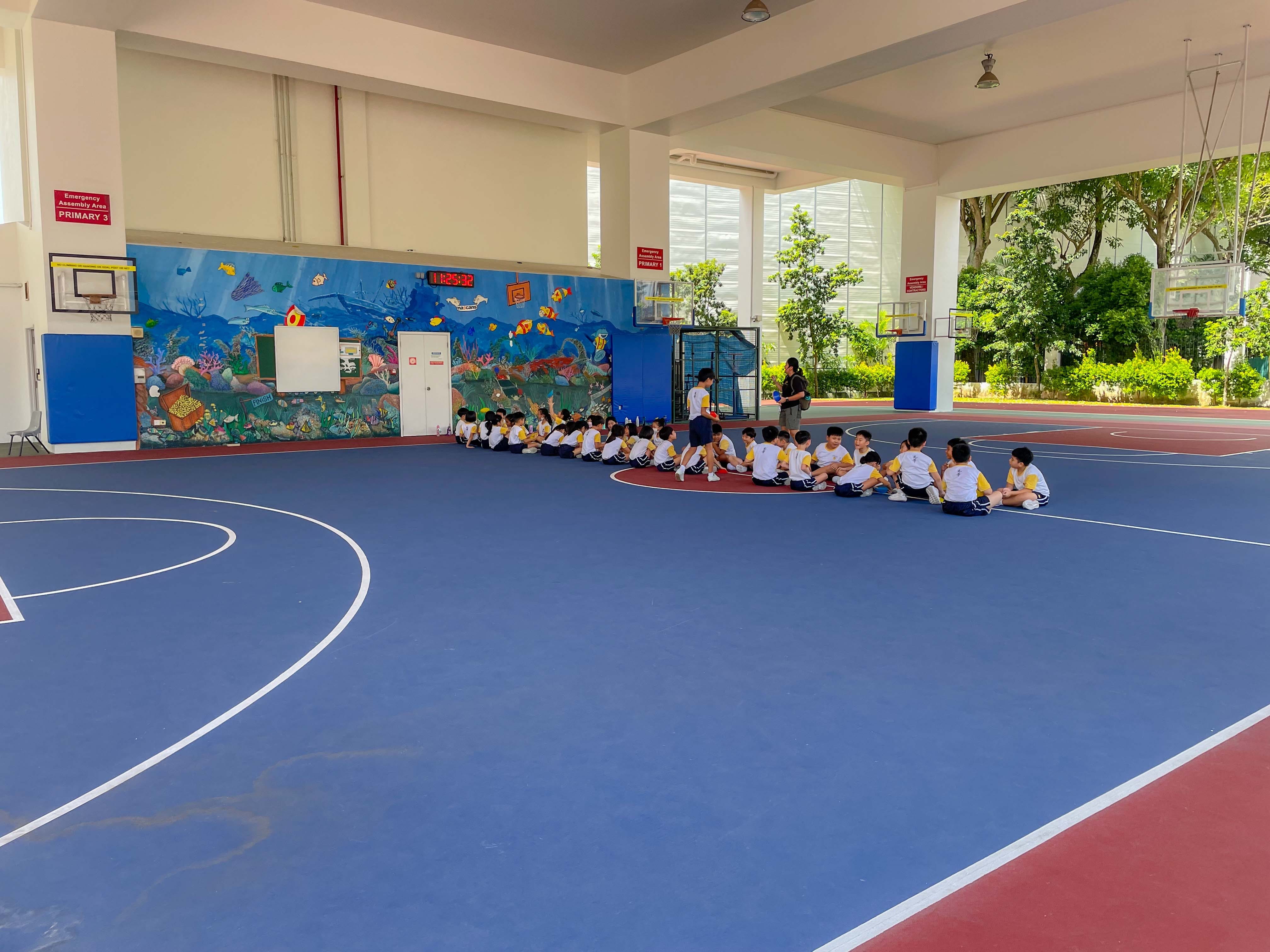 Children in white and yellow uniforms sit in a semicircle on a blue indoor basketball court with an ocean mural on the wall.