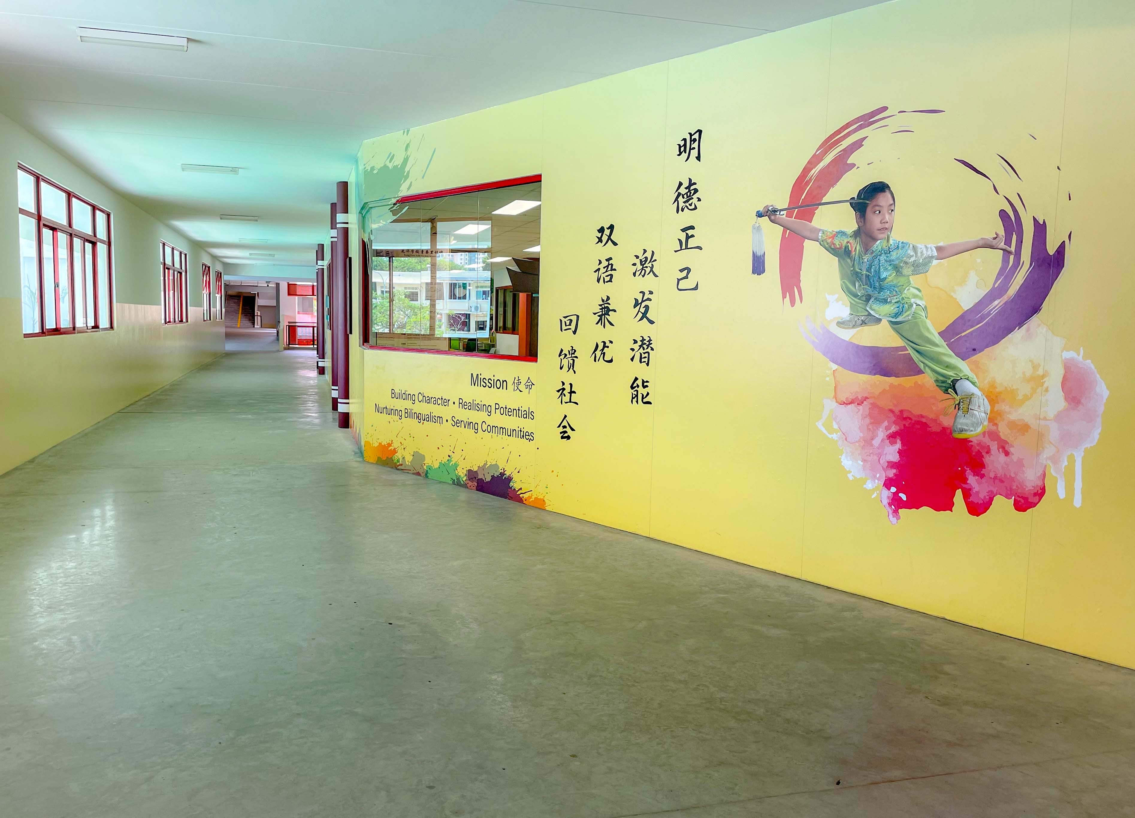 A school corridor with a yellow mural showing a girl doing martial arts, Chinese characters, and a bilingual mission statement.