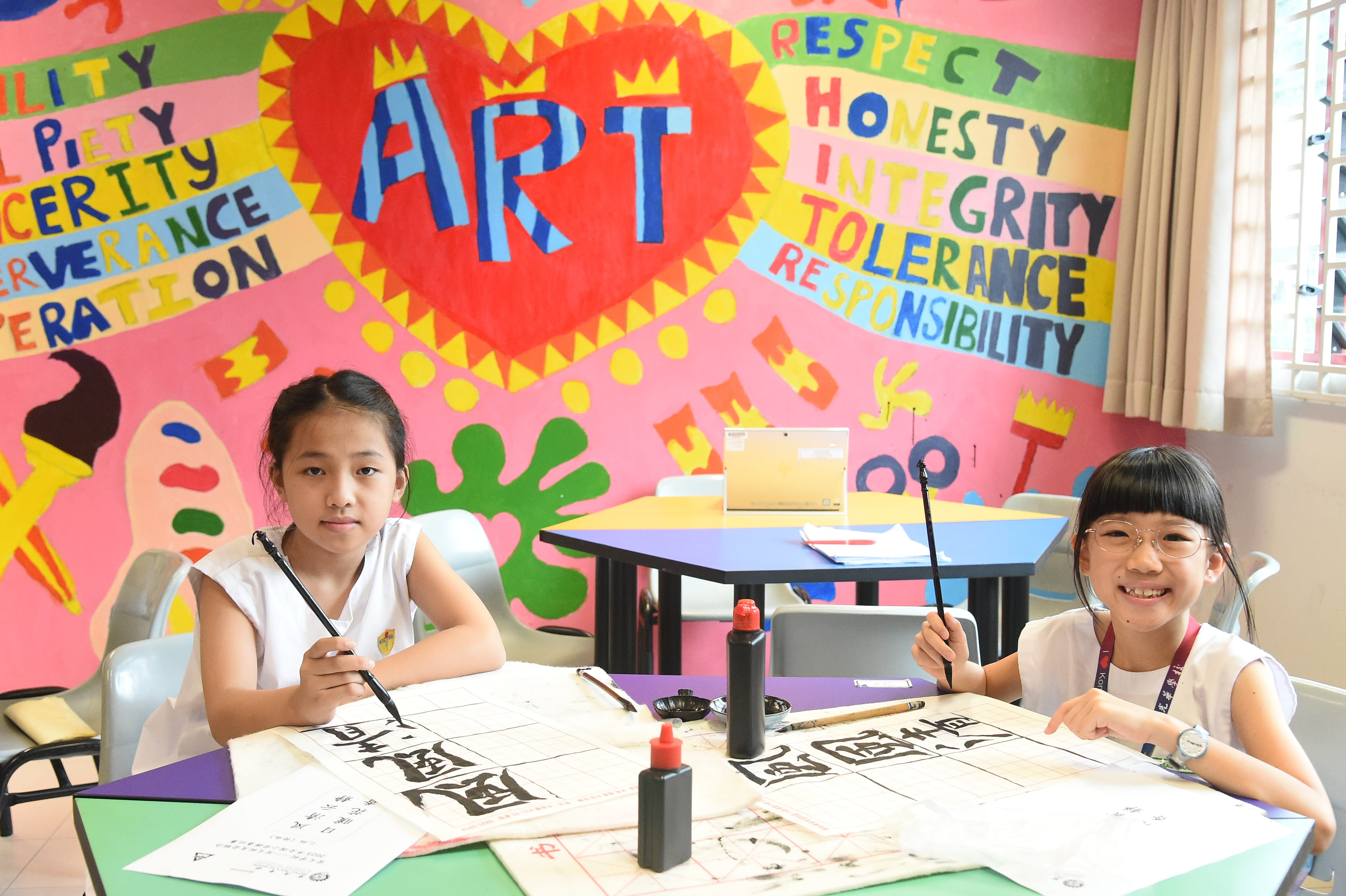 Two young girls practice Chinese calligraphy with ink brushes at a table before a colorful mural reading "ART."