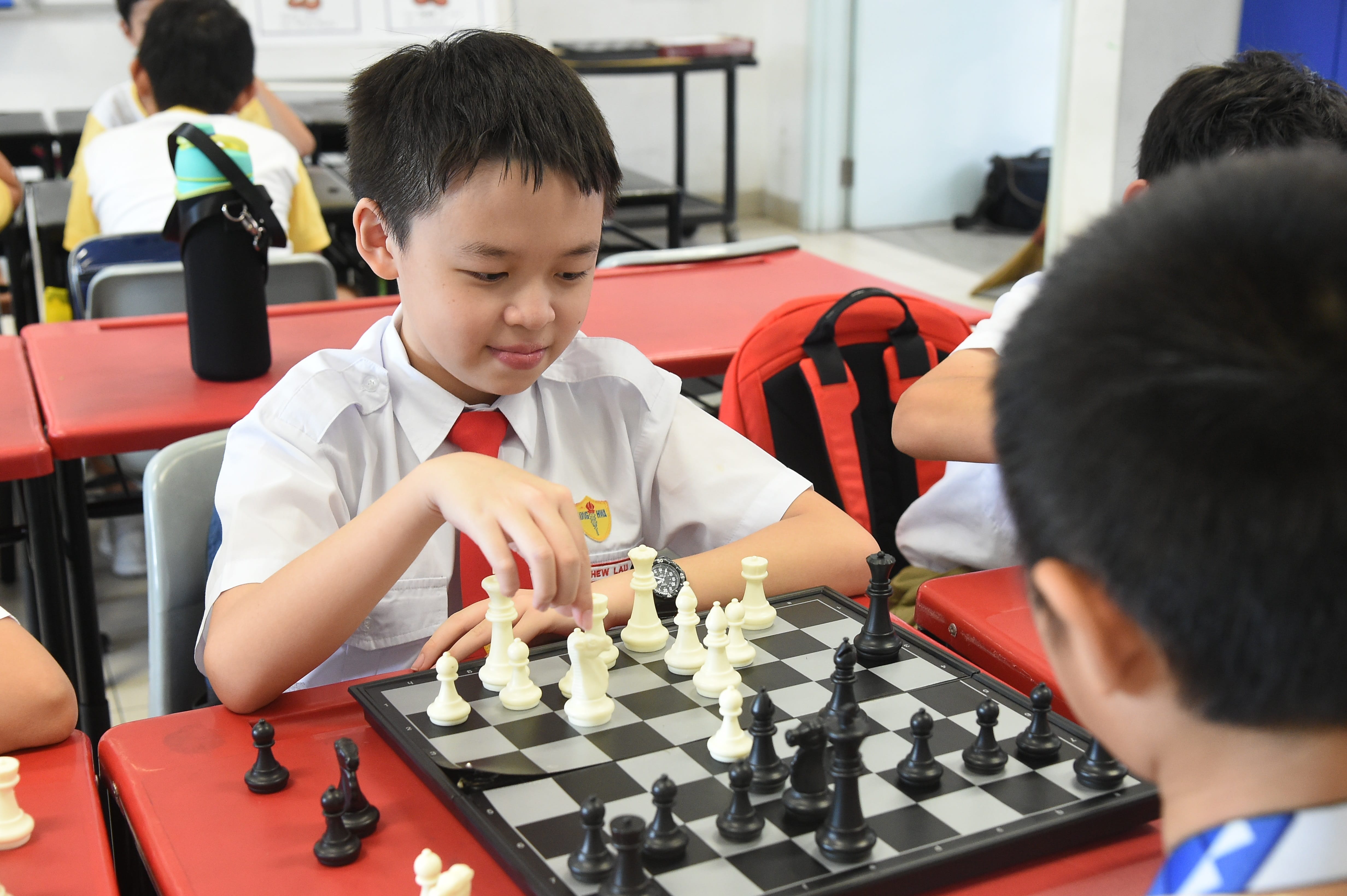 A young boy in school uniform moves a white chess piece on a board while facing an opponent across a red table.