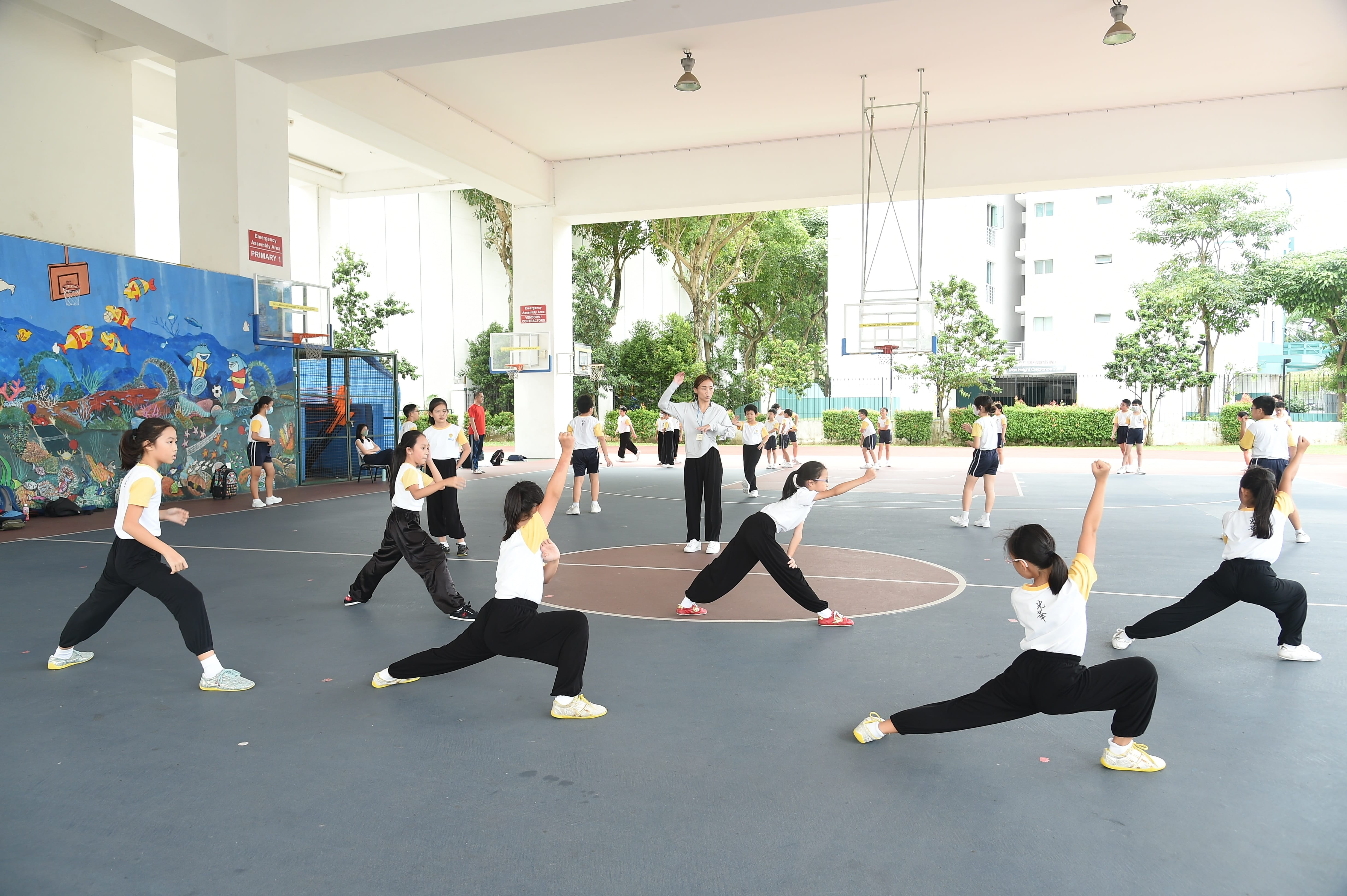 Children in school uniforms practice martial arts stances on a covered outdoor court, guided by an instructor.