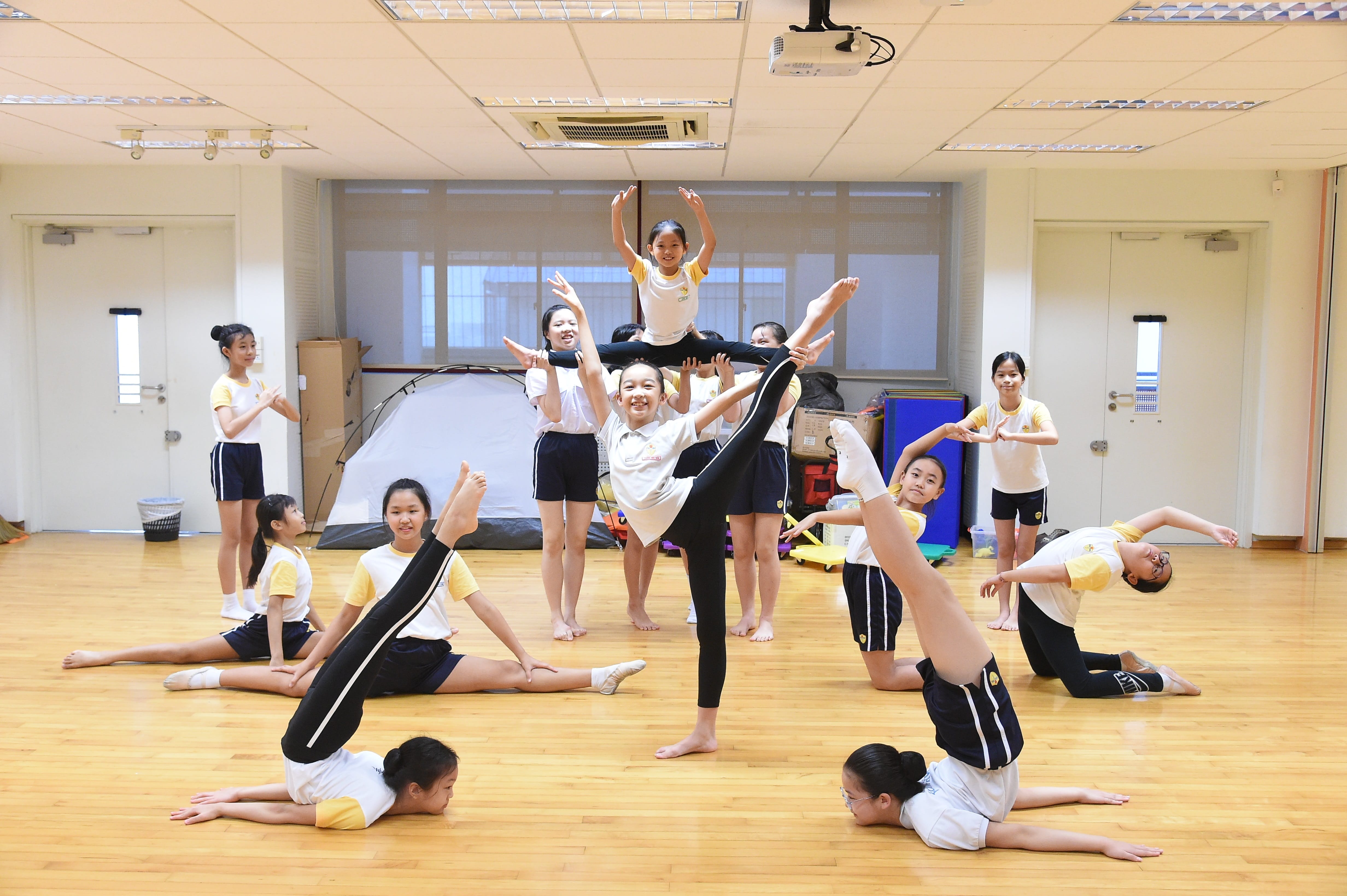A group of children in white and yellow uniforms performing acrobatic and gymnastics poses on a wooden floor indoors.