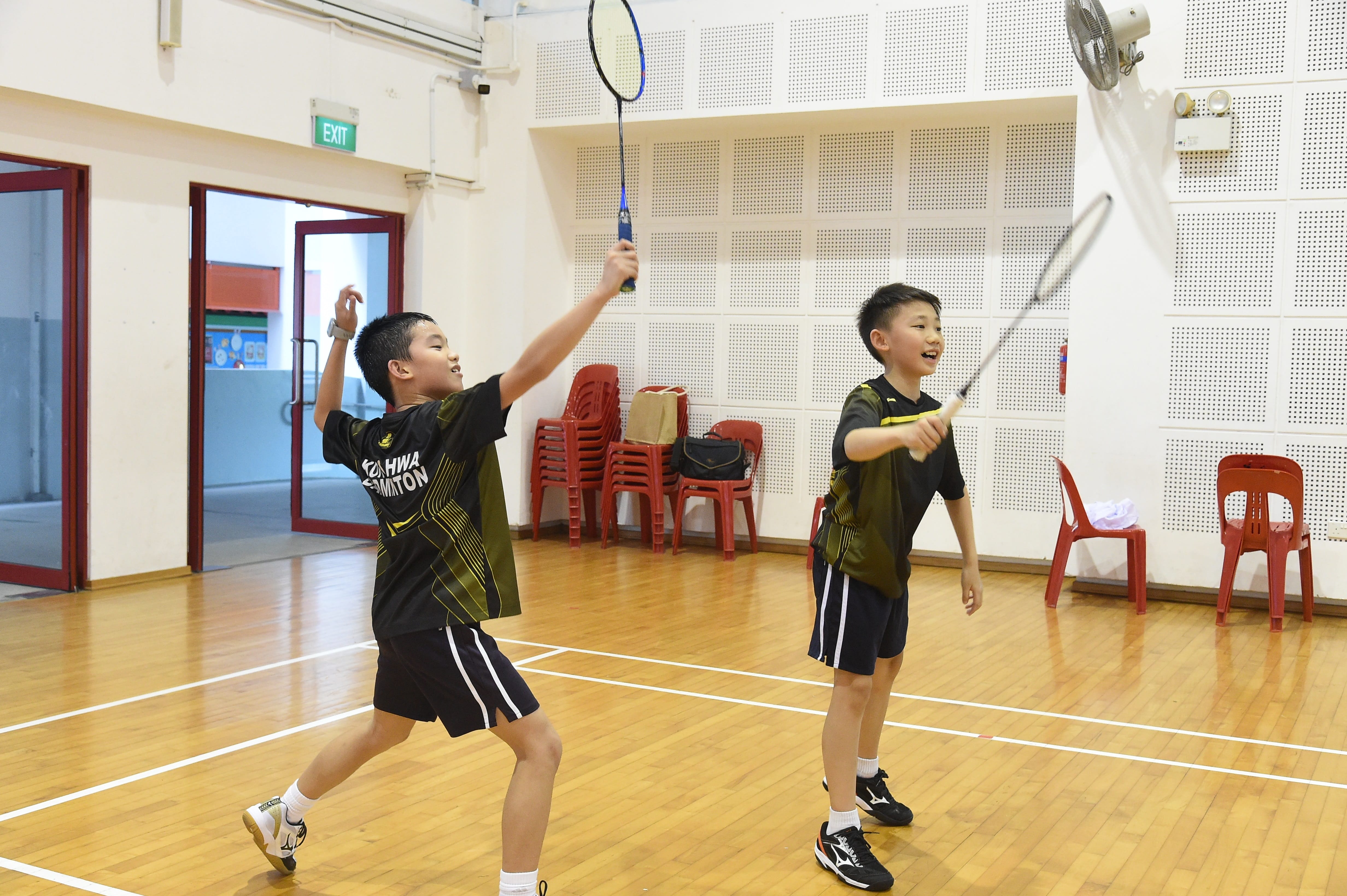 Two young boys in matching black badminton uniforms play badminton on an indoor wooden court.