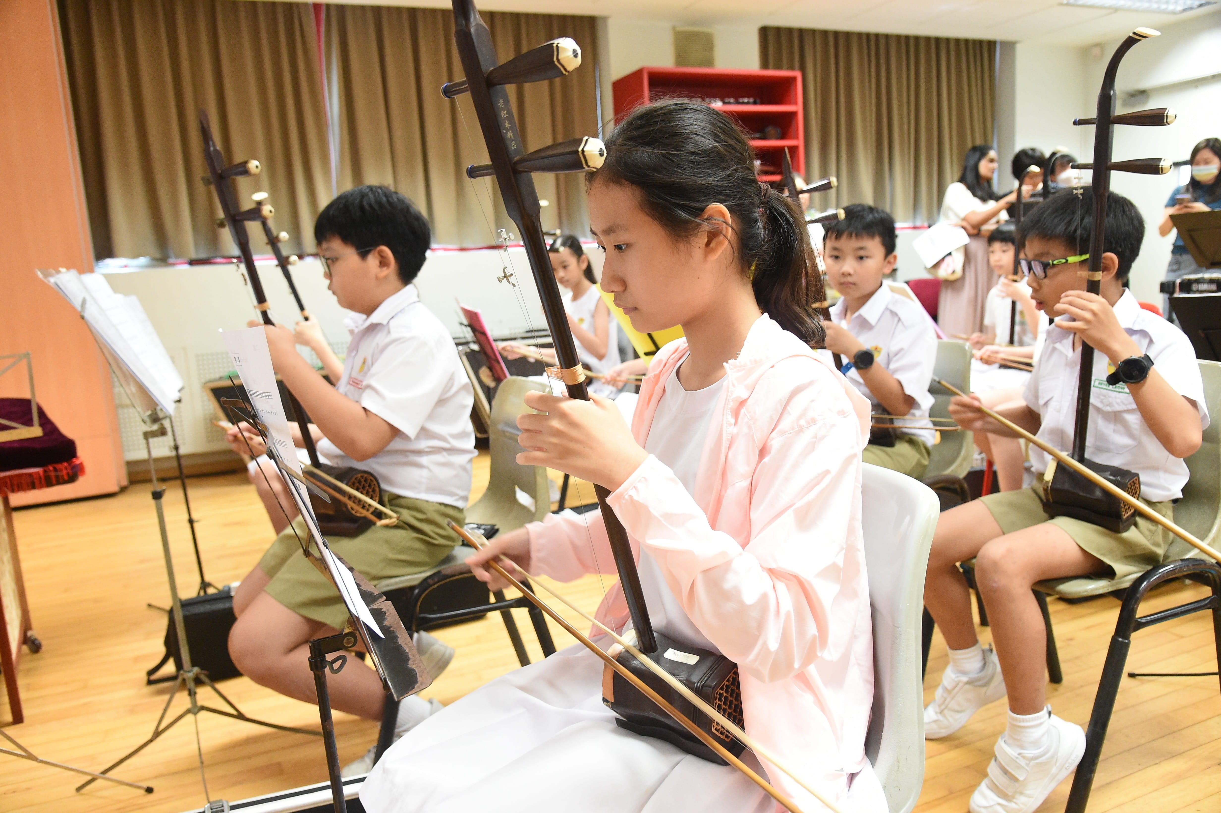 Several children in school uniforms sit in a classroom playing erhu instruments while reading sheet music stands.