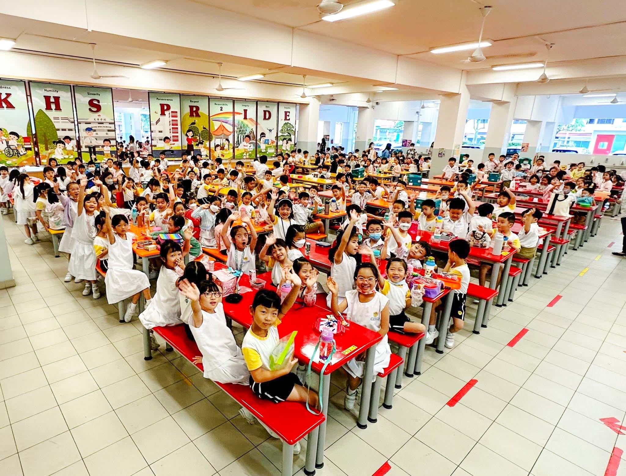 Large group of young school children in white uniforms sitting at red cafeteria tables, waving and smiling at the camera.
