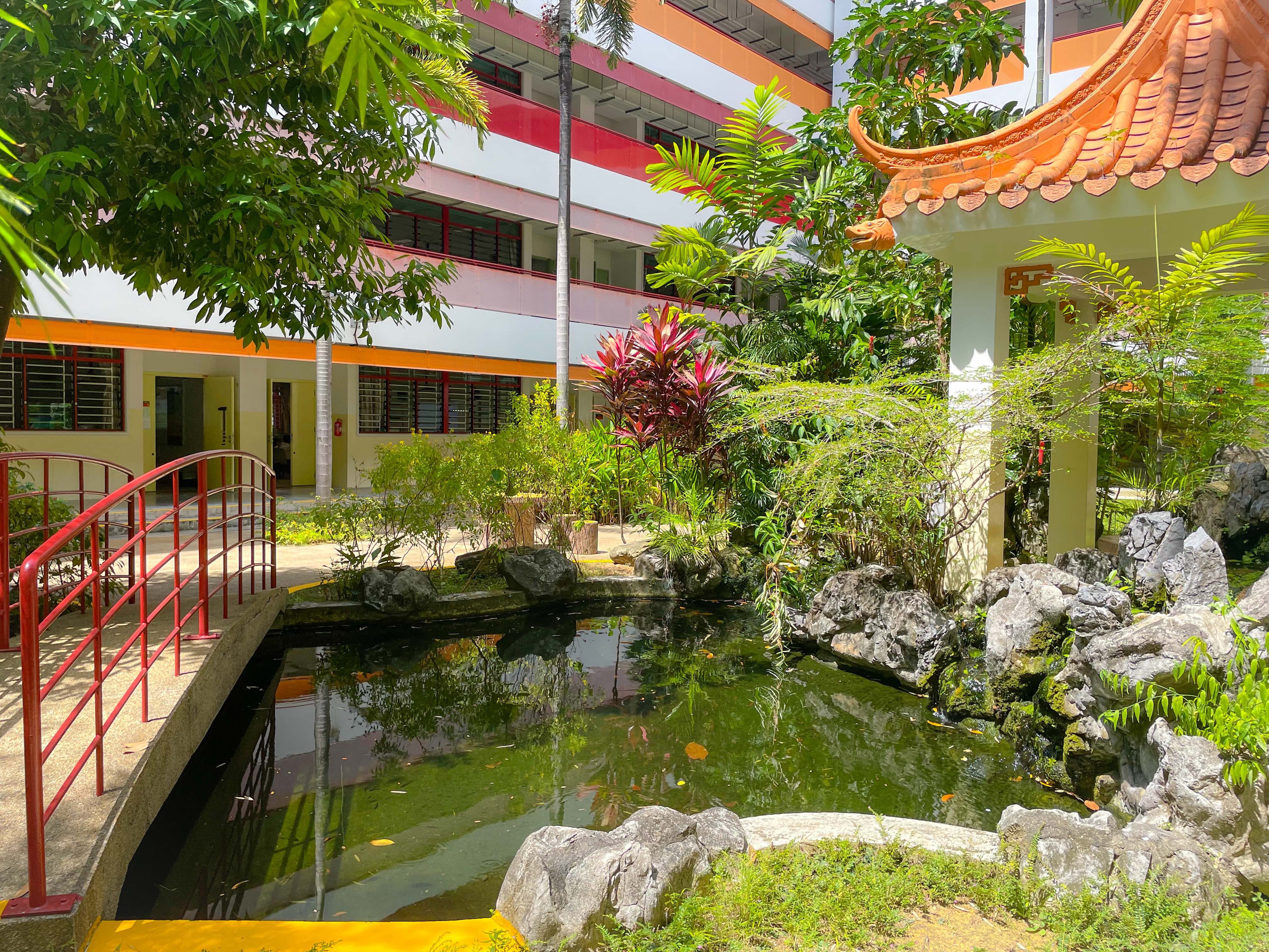 A pond with rocks and lush plants beside a red arched bridge and a Chinese-style pavilion near a colorful building.
