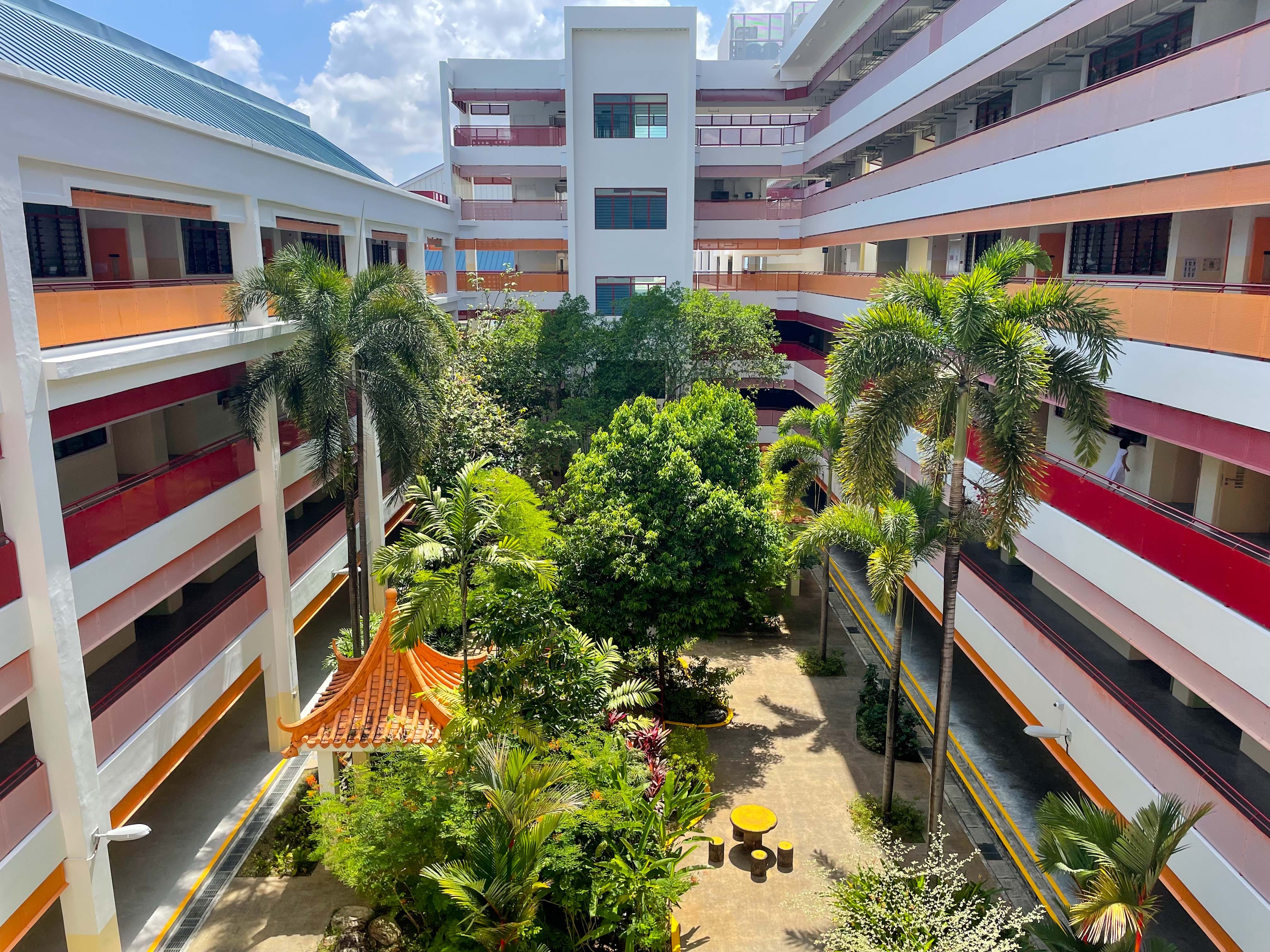 Aerial view of a multi-story school courtyard with palm trees, a small pagoda, and red-and-white corridor buildings.