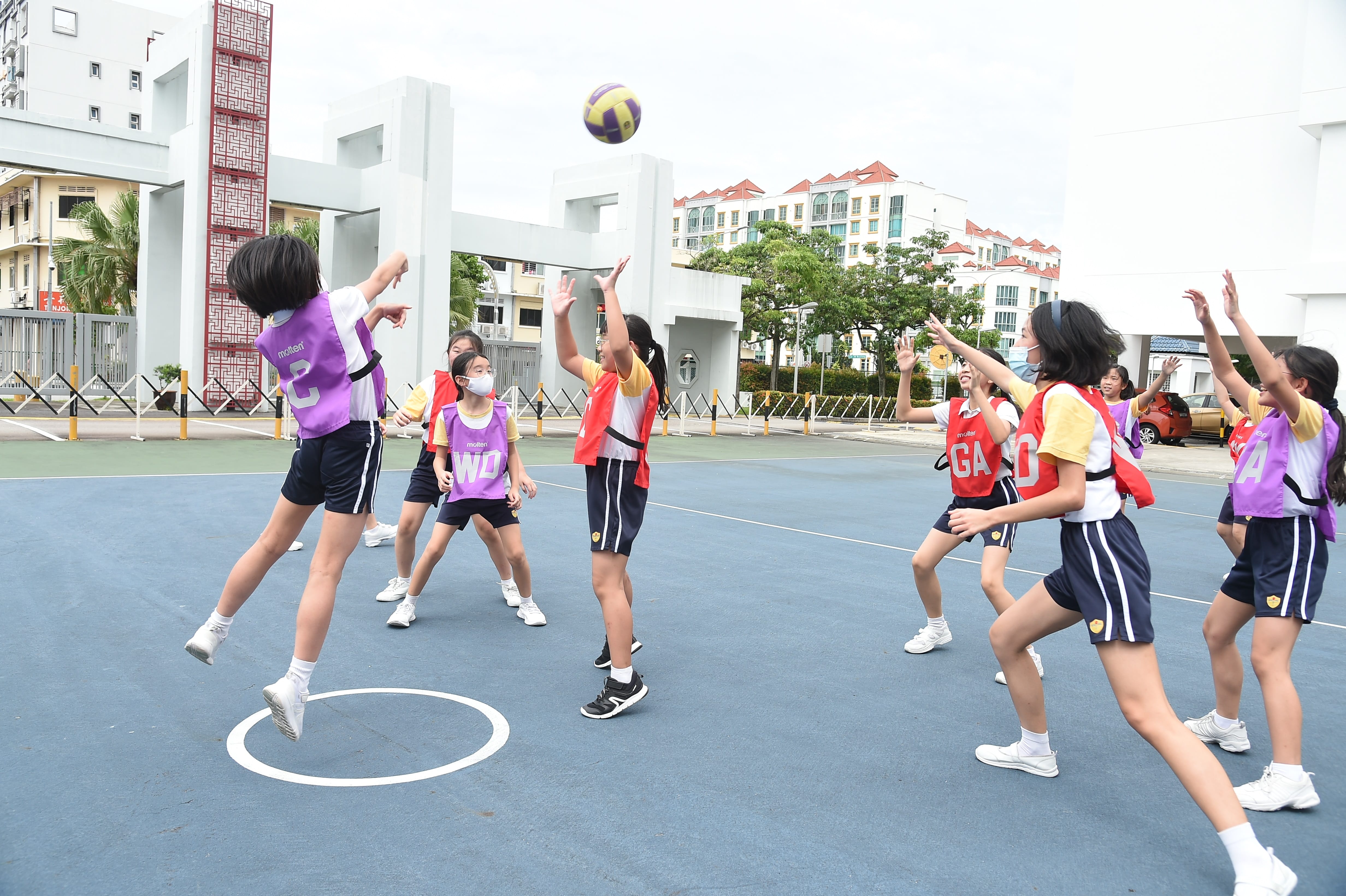 Girls in red and purple bibs play netball on an outdoor court, with one jumping to intercept a ball in the air.