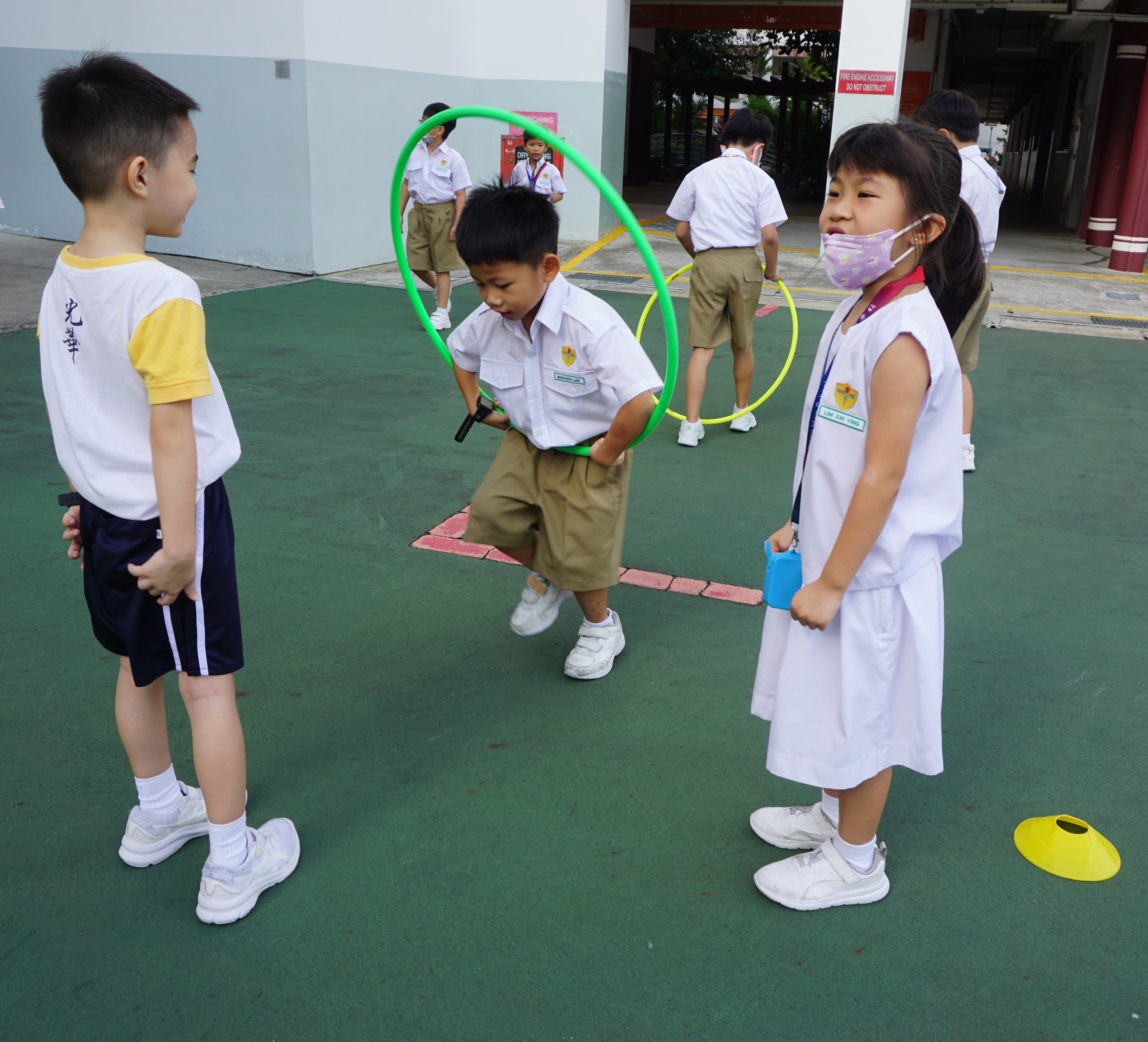 Young school children in uniforms play with green and yellow hula hoops on an outdoor court.