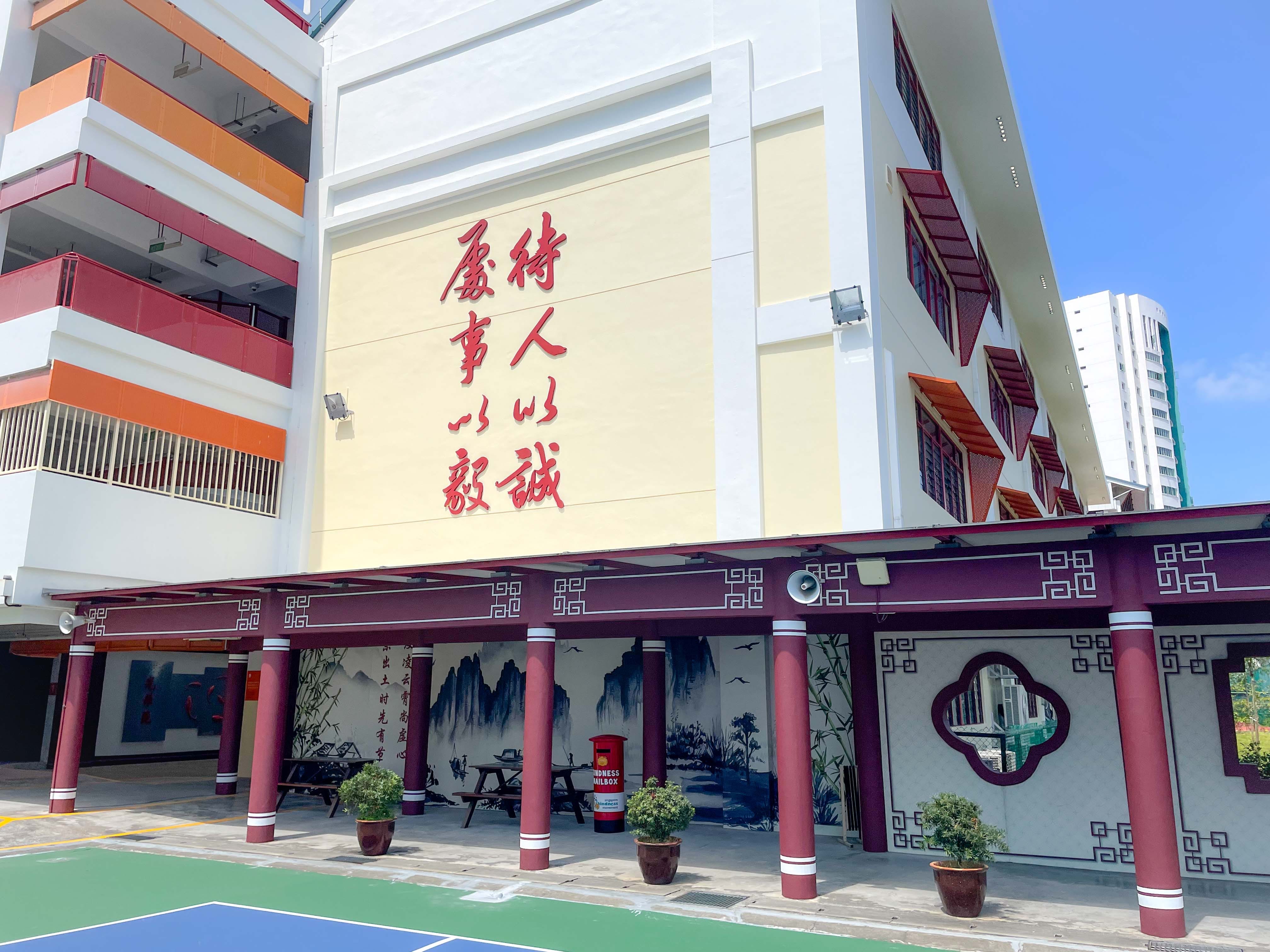 A Chinese-style school building with red columns, landscape murals, and large red Chinese characters on a cream wall.