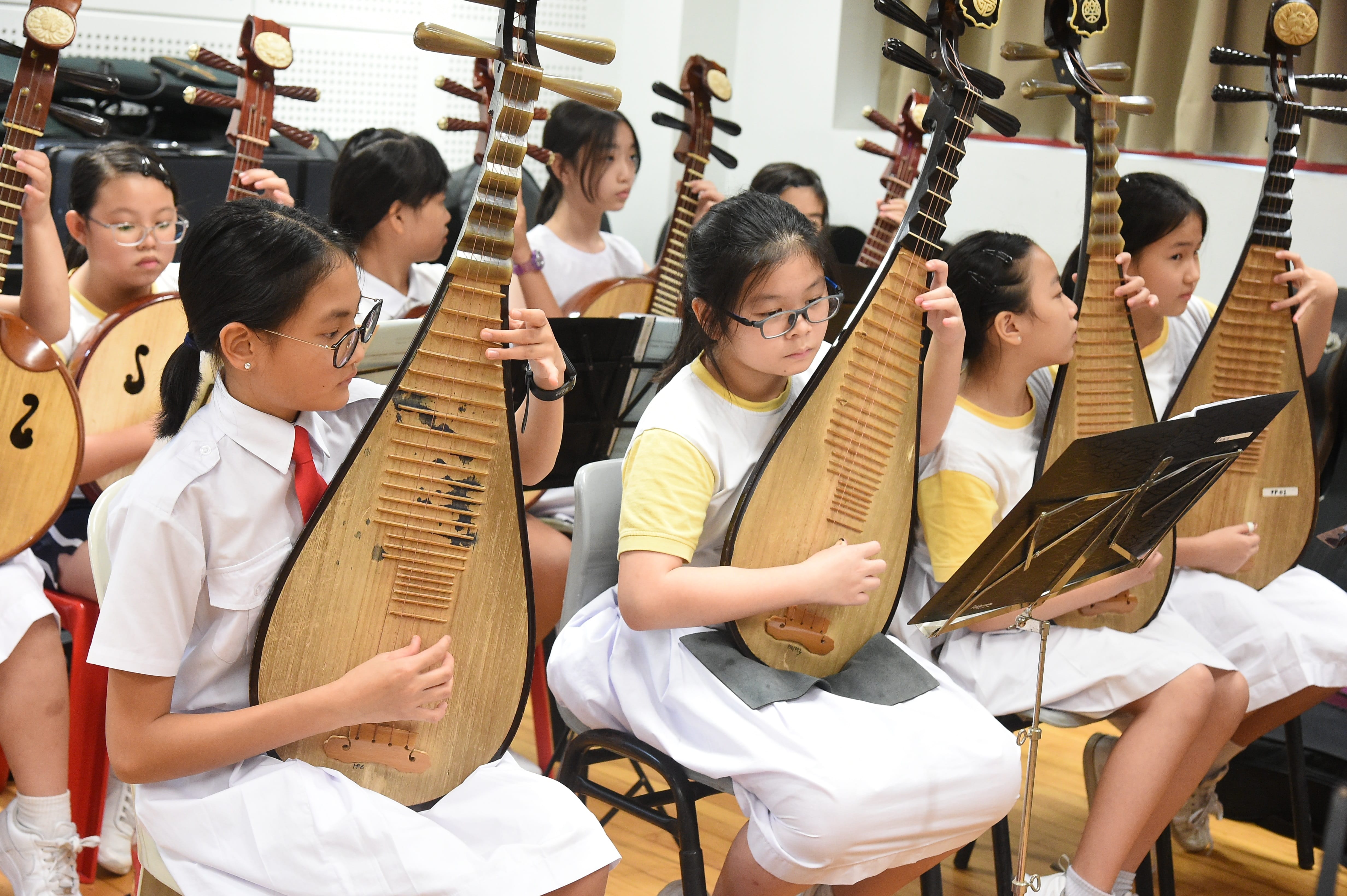 A group of young students in white uniforms playing pipa, a Chinese lute, during a music rehearsal.