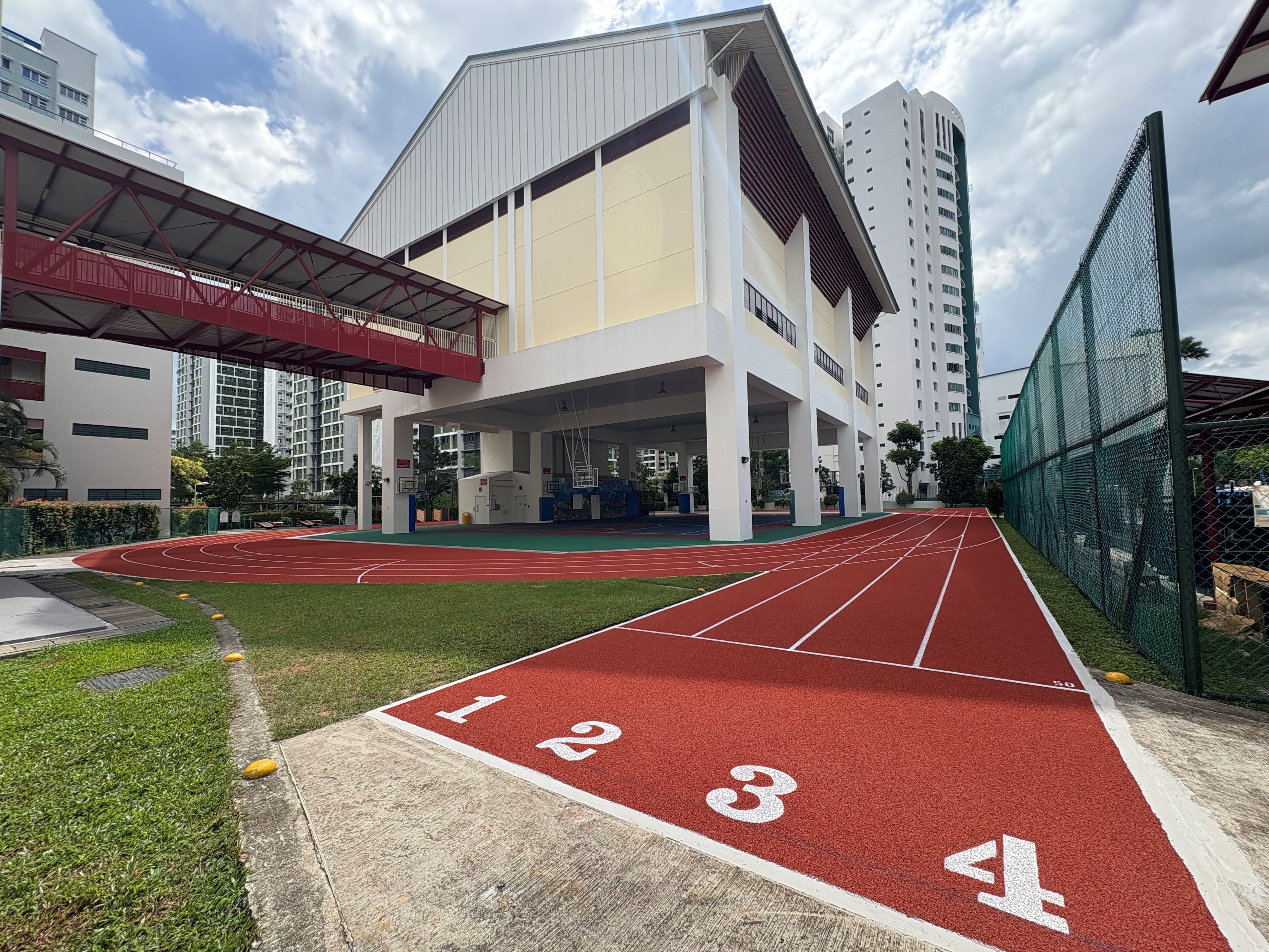 Red running track with lanes numbered 1 to 4 beside a school building and tall residential towers under a cloudy sky.