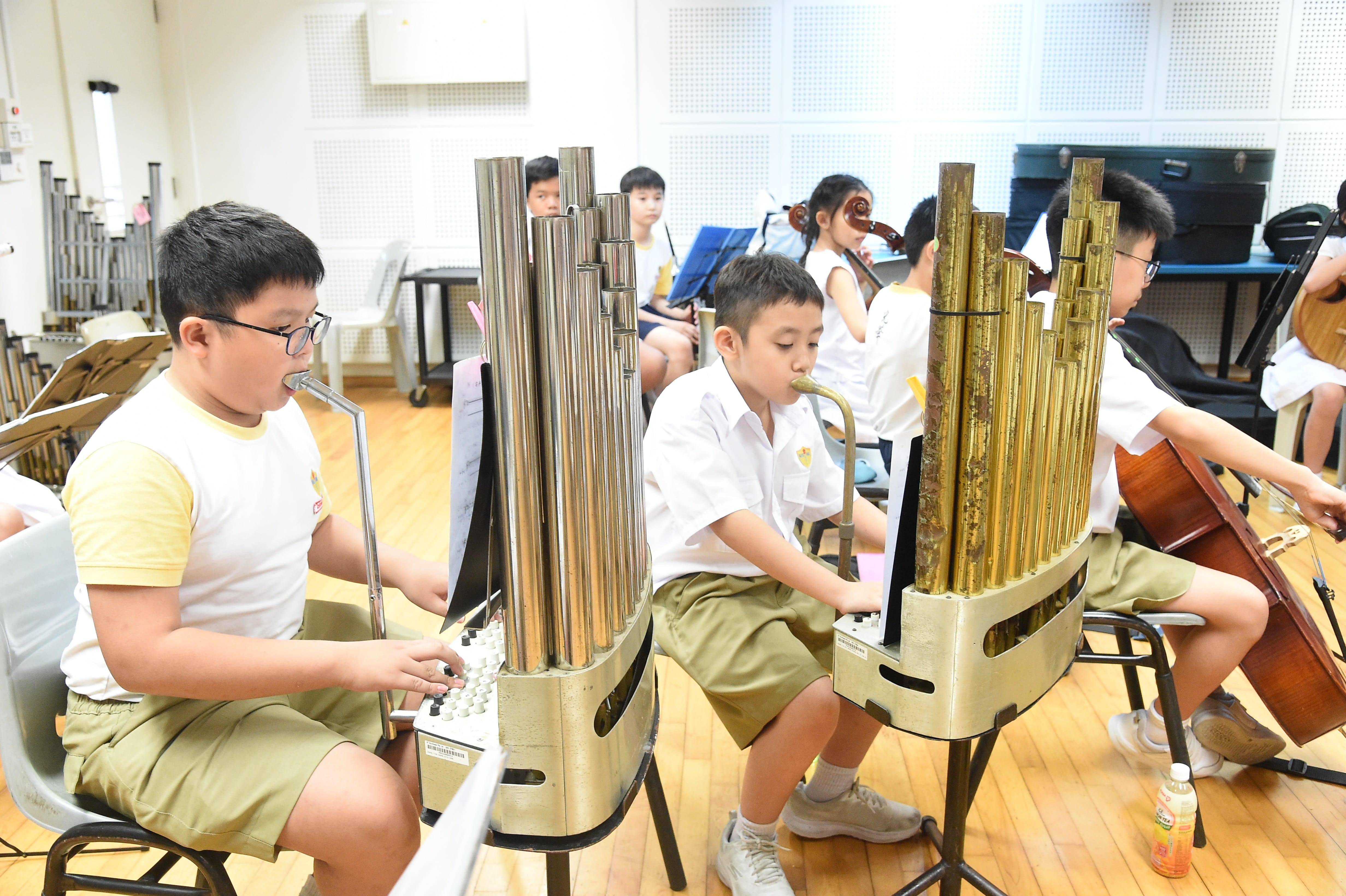 Children in school uniforms play pipe organs and string instruments during a music rehearsal in a classroom.