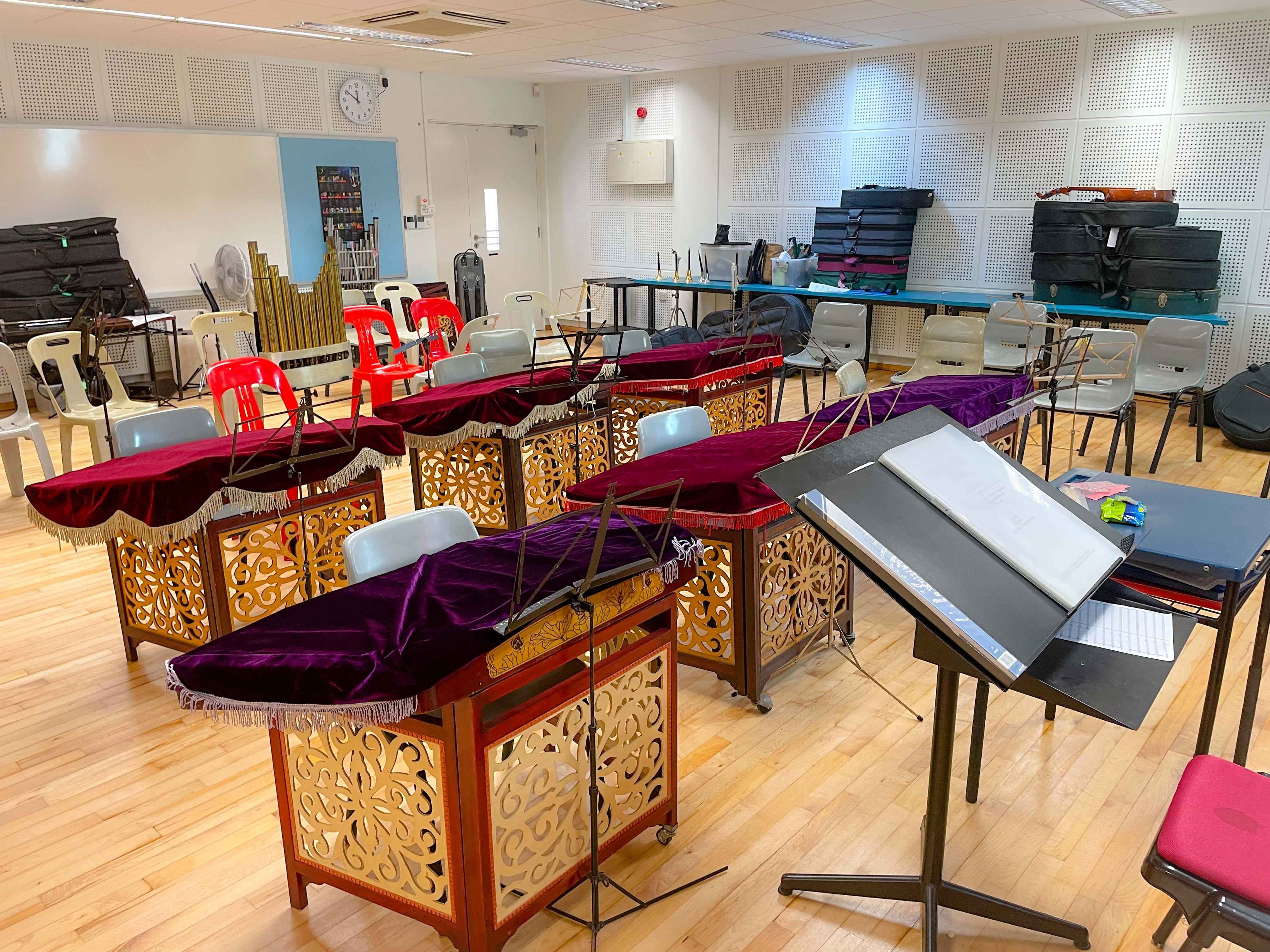 A rehearsal room with gamelan instruments covered in red and purple velvet, music stands, and chairs on a wooden floor.