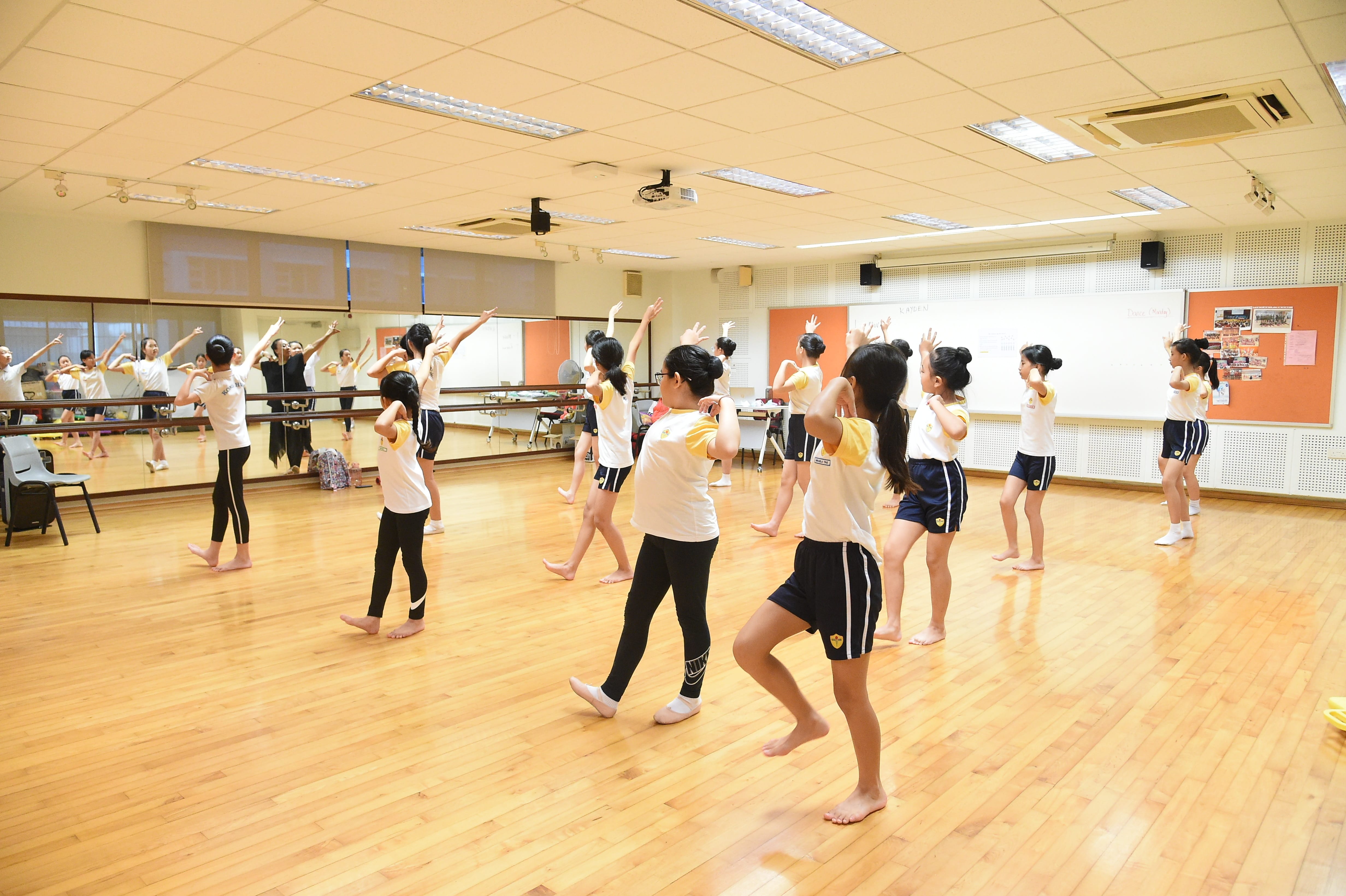 Children in white shirts practice dance moves barefoot in a mirrored studio with wooden floors.