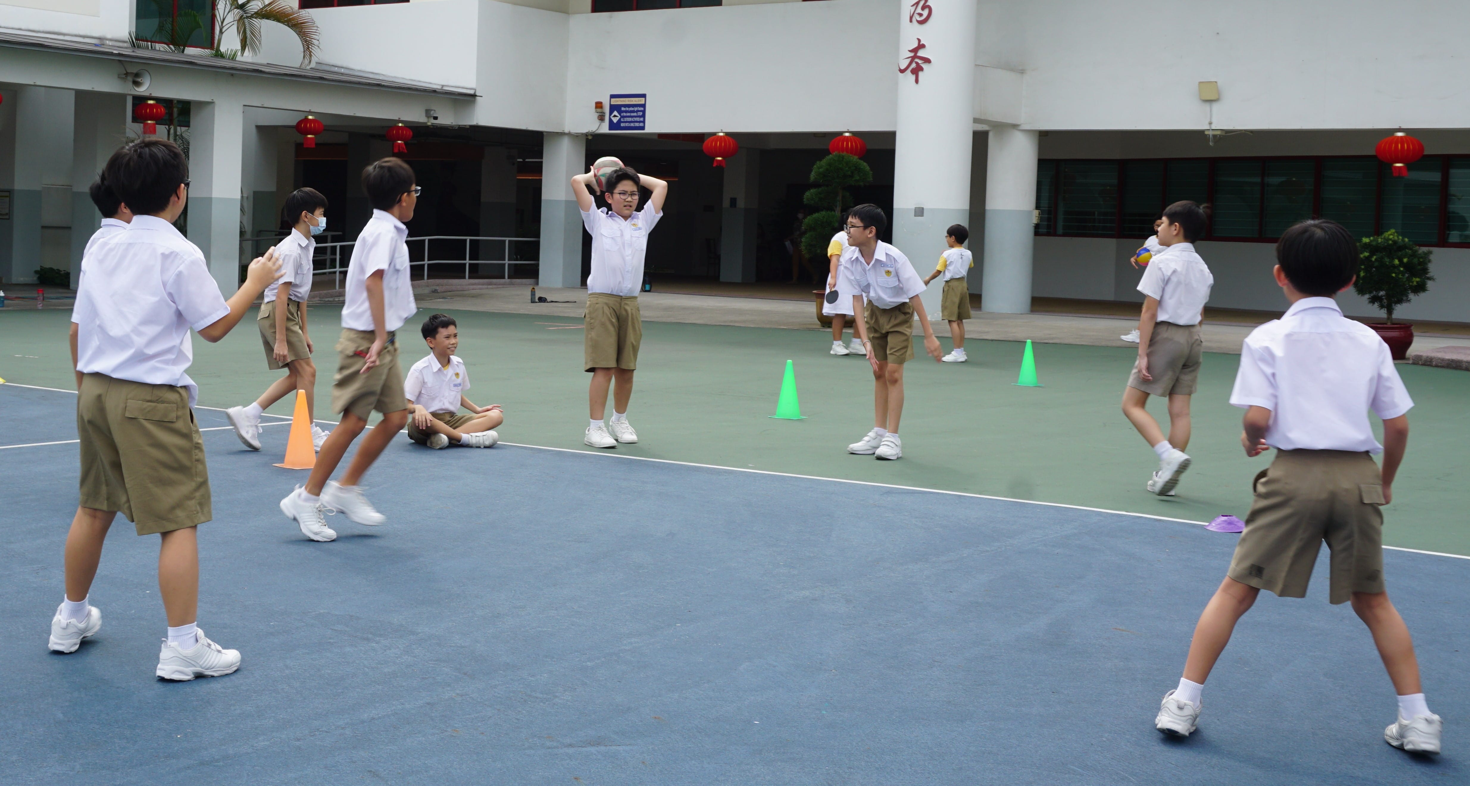 Students in white shirts and khaki shorts play a ball game on an outdoor school court with colored cones.