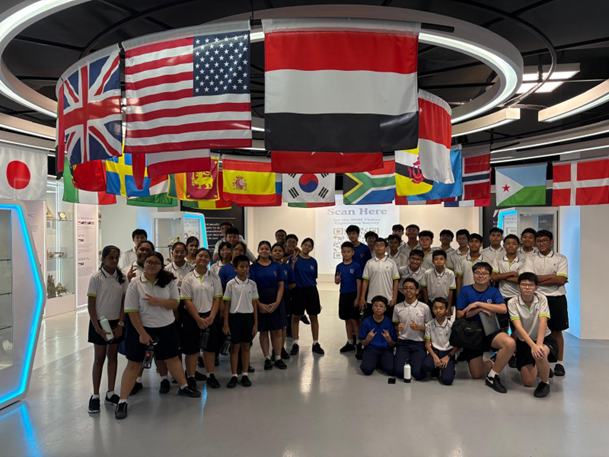 A group of students in uniforms pose indoors beneath a circular display of international flags hanging from the ceiling.