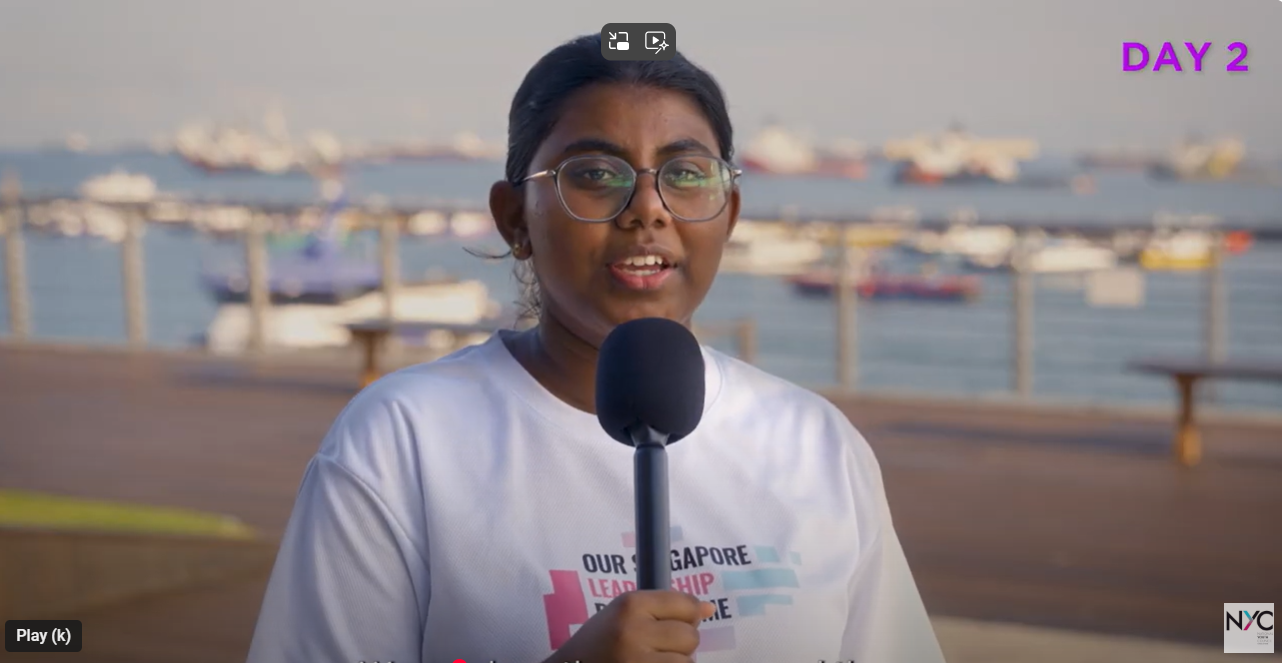 Young woman with glasses holding a microphone outdoors near a waterfront with boats, wearing a Singapore Leadership shirt.