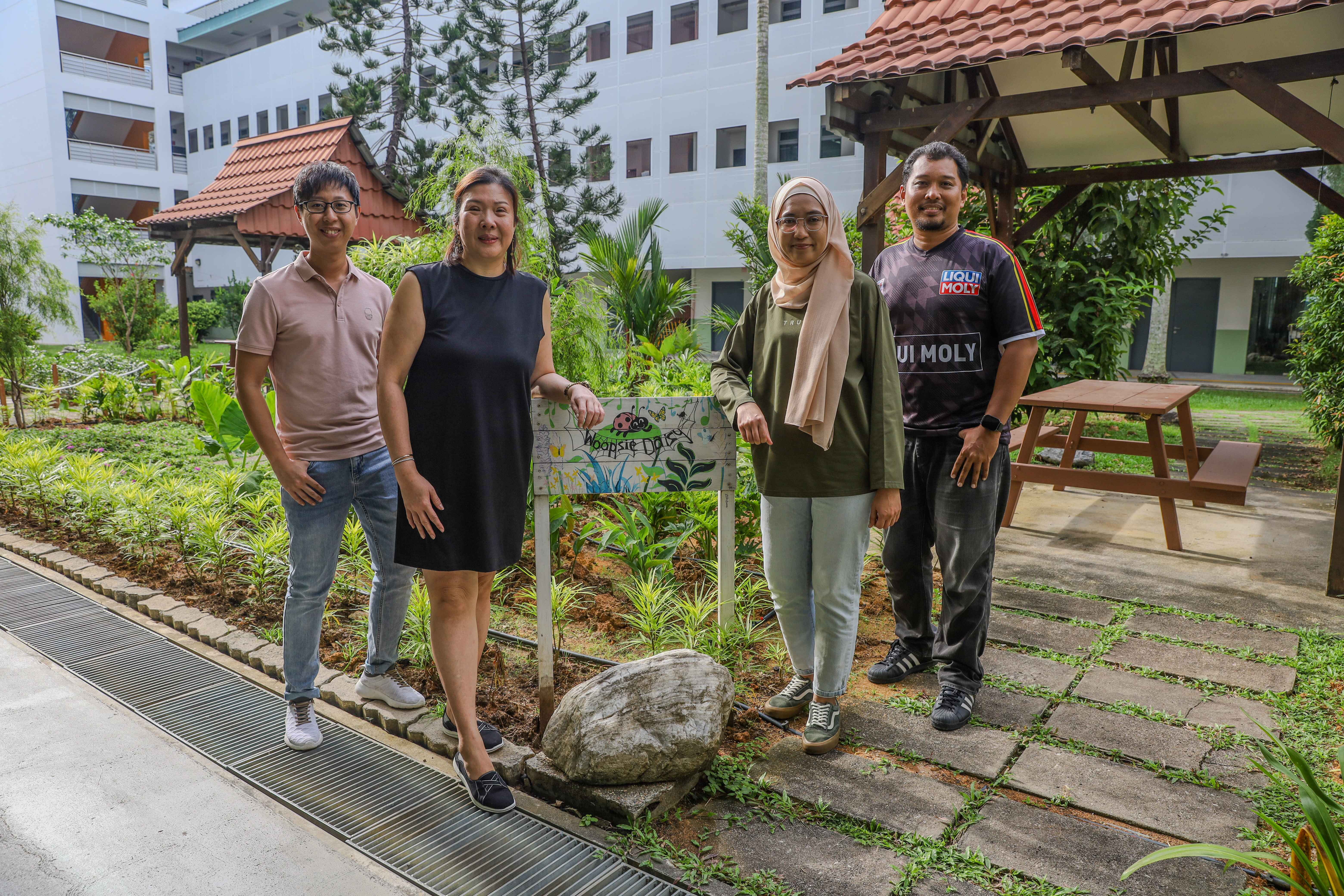 Four people stand smiling beside a painted garden sign in a green courtyard with white buildings behind them.