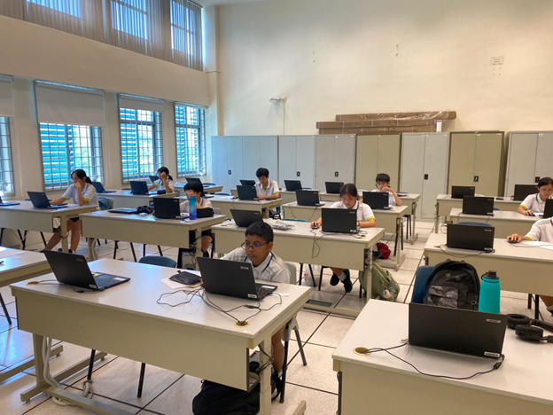 Students in school uniforms sit at individual desks with laptops in a bright classroom.