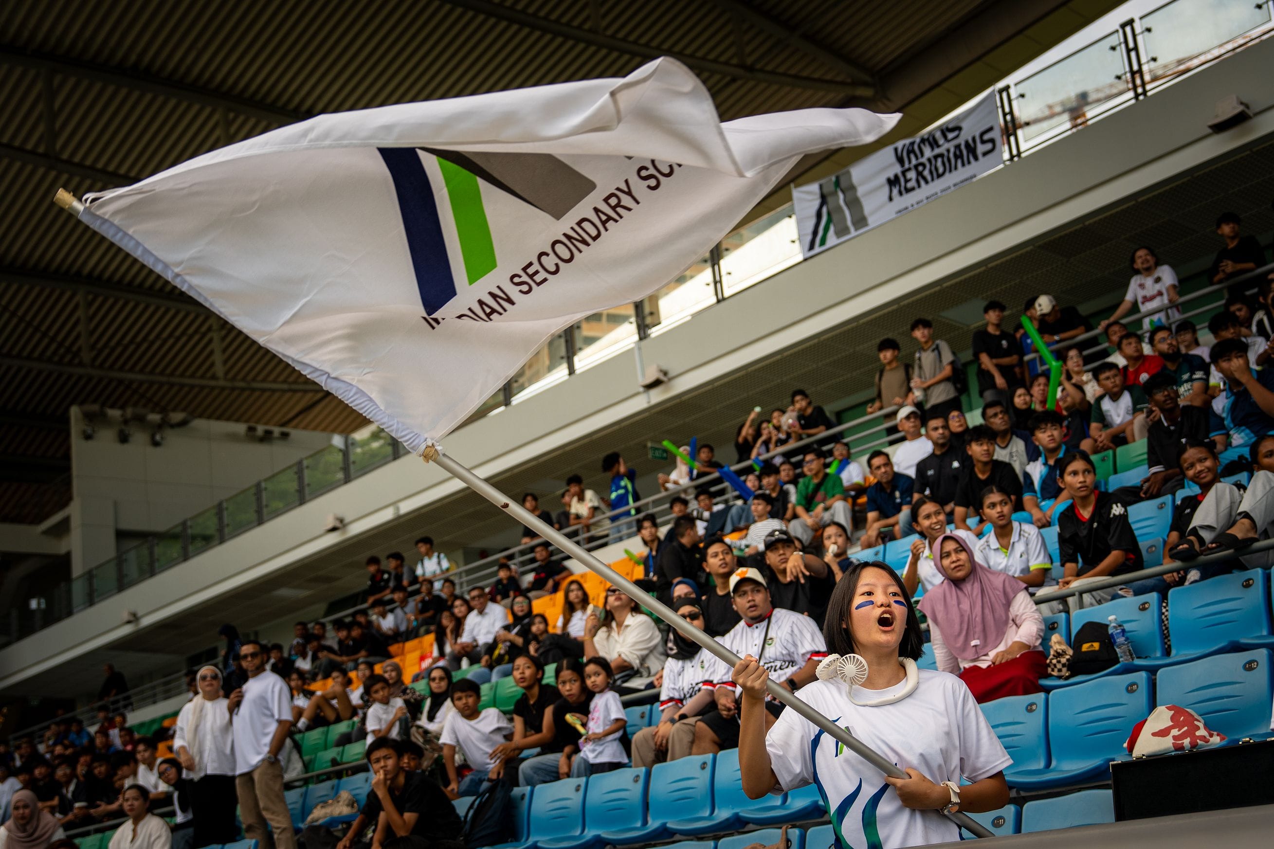 A girl with face paint waves a large white school flag in a packed stadium with cheering spectators behind her.