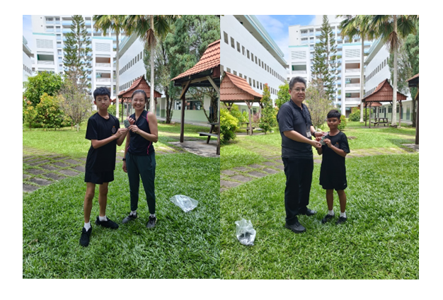 Two pairs of people in black clothing stand on grass outside a building with gazebos, exchanging small items.