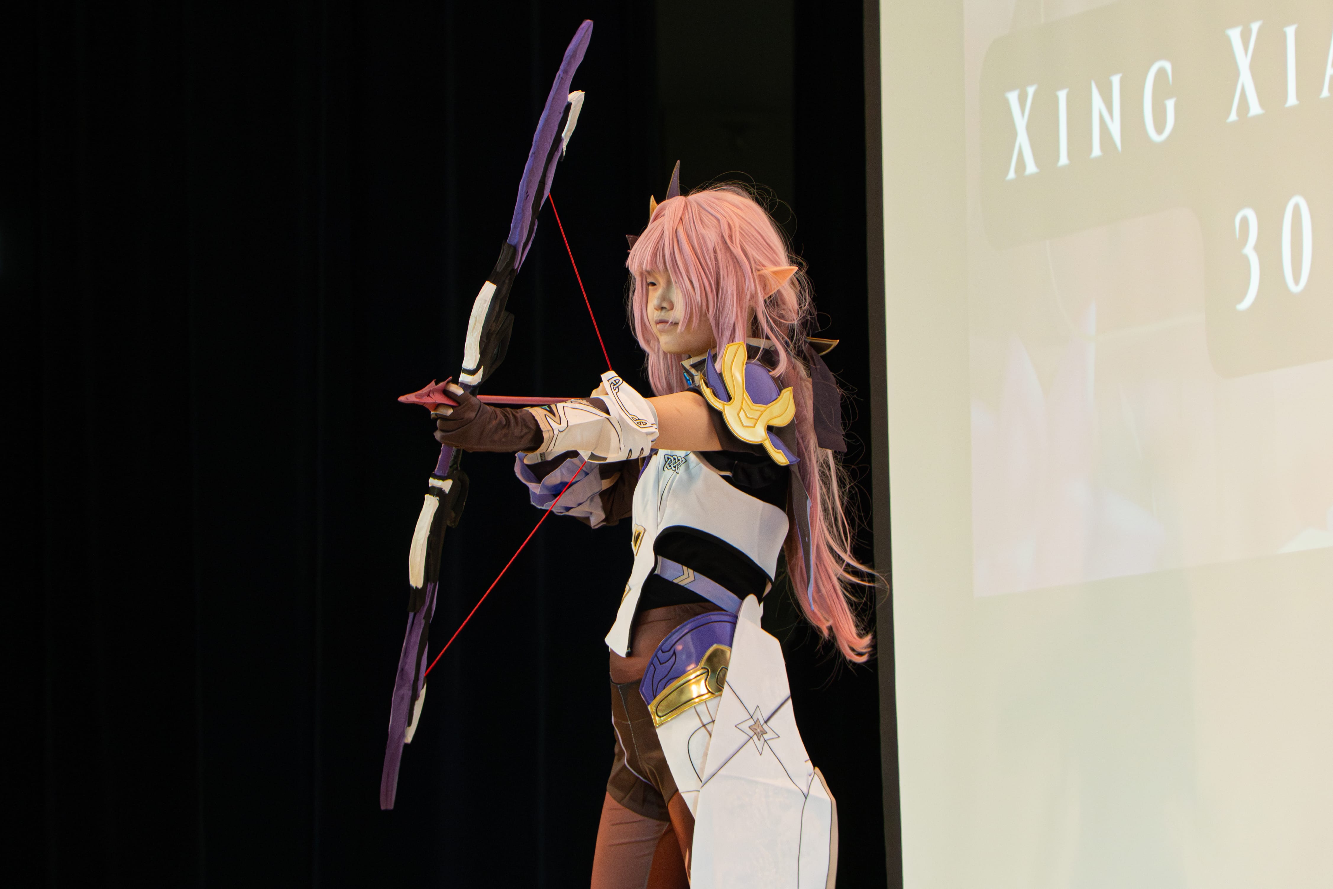Person in anime cosplay with pink wig and fantasy armor poses aiming a purple bow on a stage.
