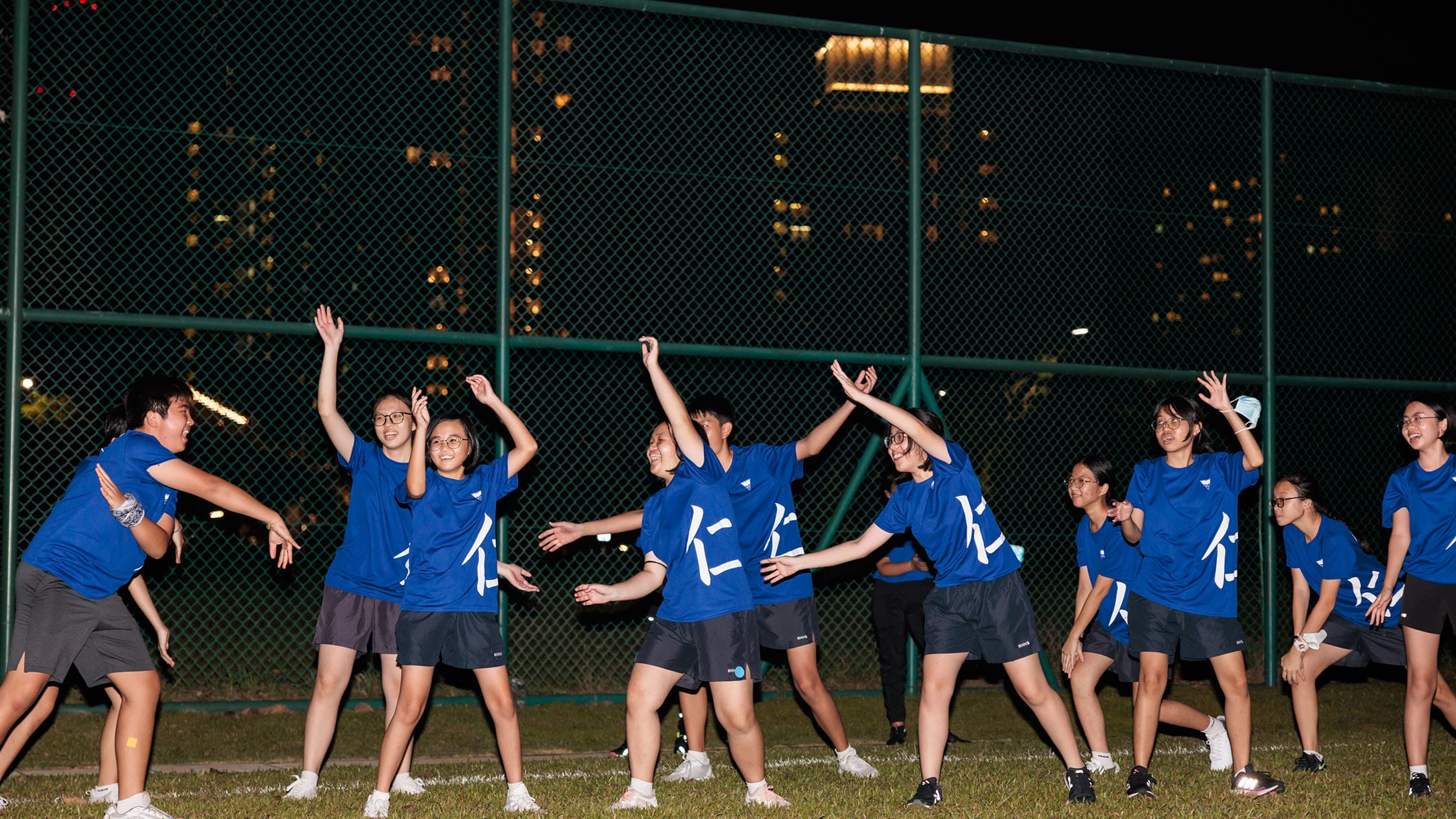 A group of youths in matching blue T-shirts with Chinese characters dance outdoors at night near a chain-link fence.
