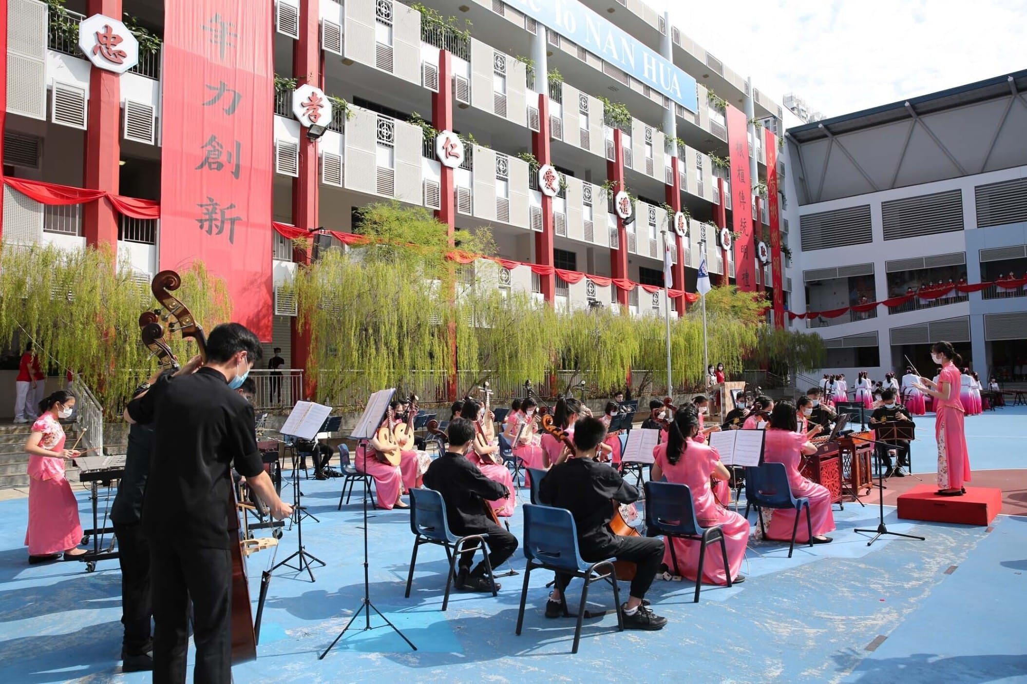 Students in pink dresses perform a Chinese orchestra outdoors at Nan Hua school, decorated with red banners bearing Chinese characters.