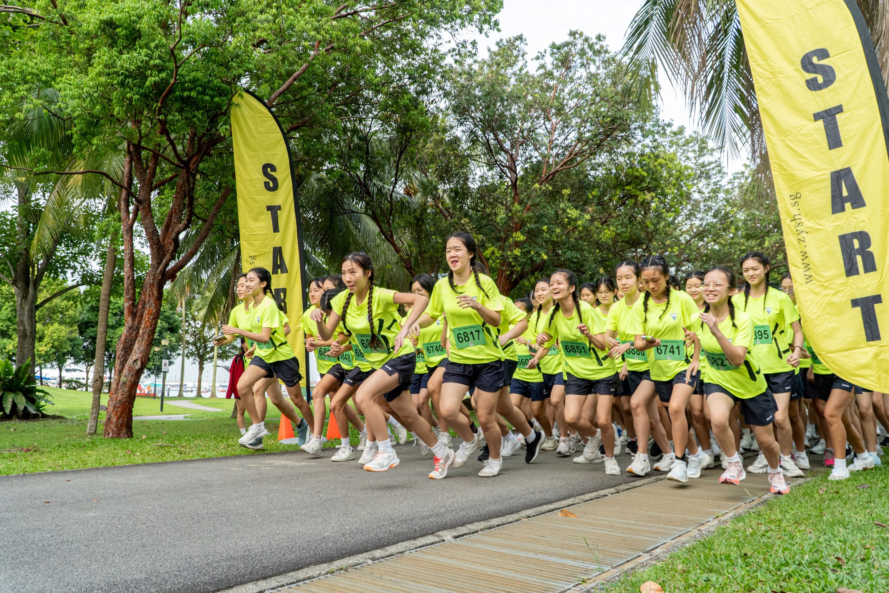 A large group of young women in yellow shirts with race bibs sprint from a start line marked by yellow banners.