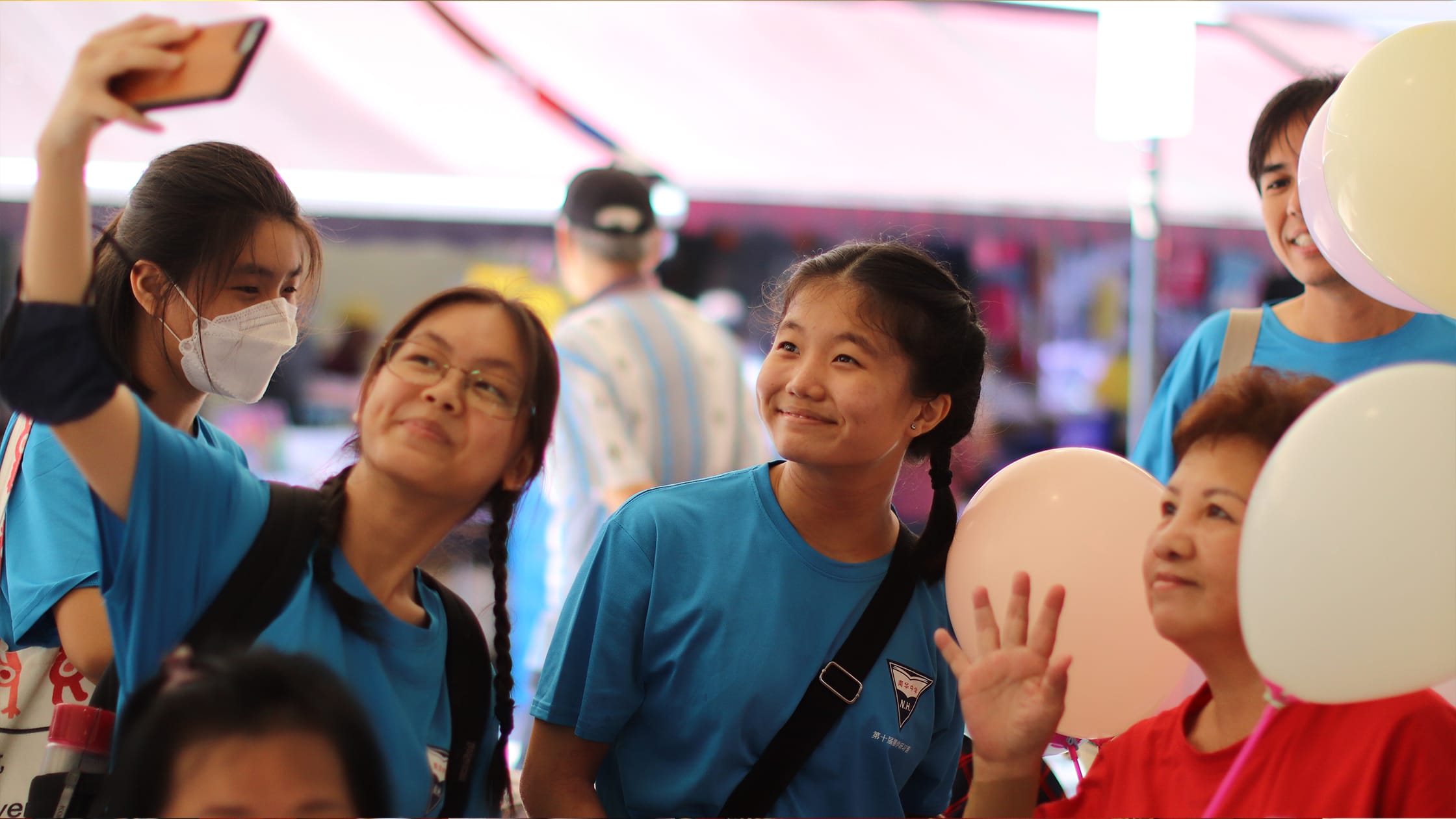 A group of young people in blue shirts at an event, one taking a selfie, another smiling, with white balloons nearby.