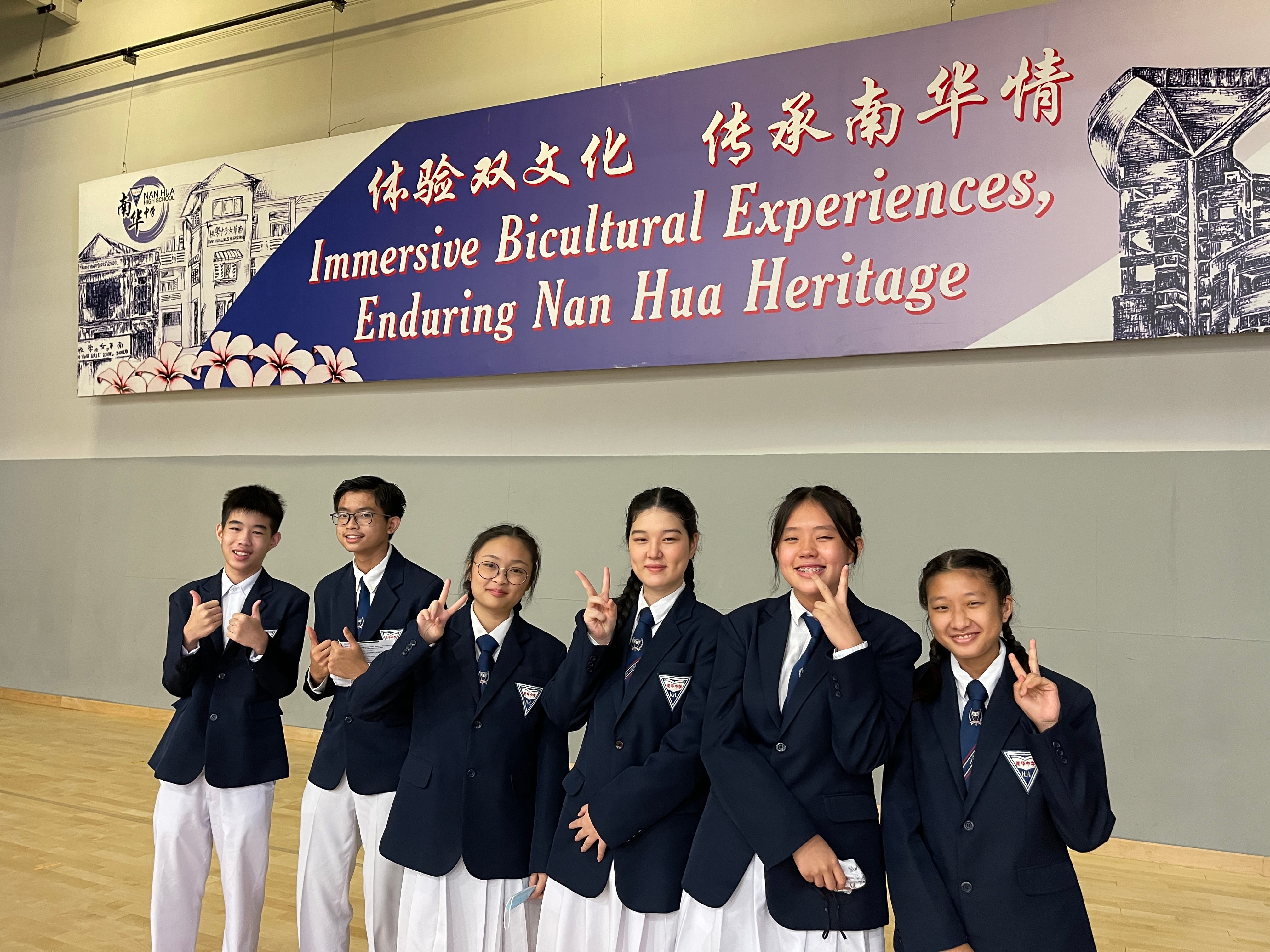 Six students in navy blazers pose with peace signs before a Nan Hua Heritage banner with Chinese and English text.