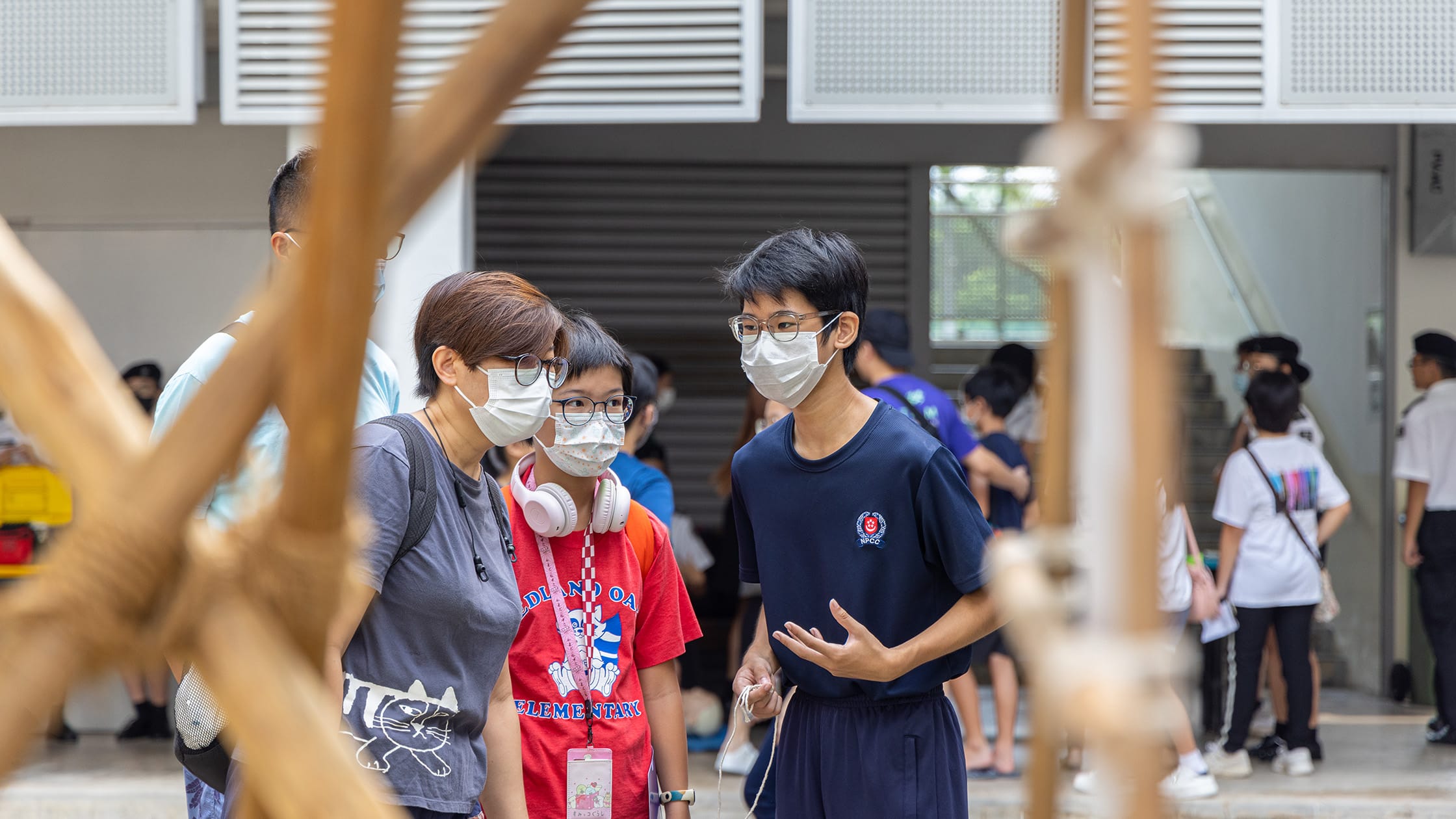 Three people wearing face masks and glasses stand talking indoors, with a crowd visible in the background.