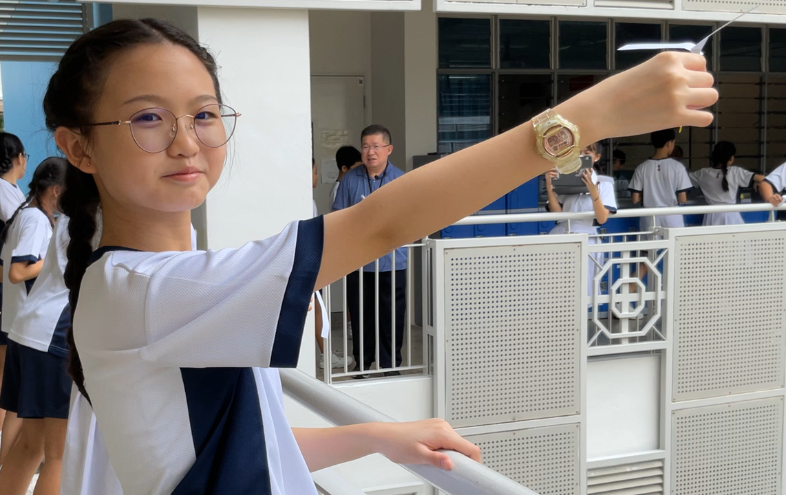 A young girl in school uniform with glasses raises her arm showing a gold digital watch on a school balcony.