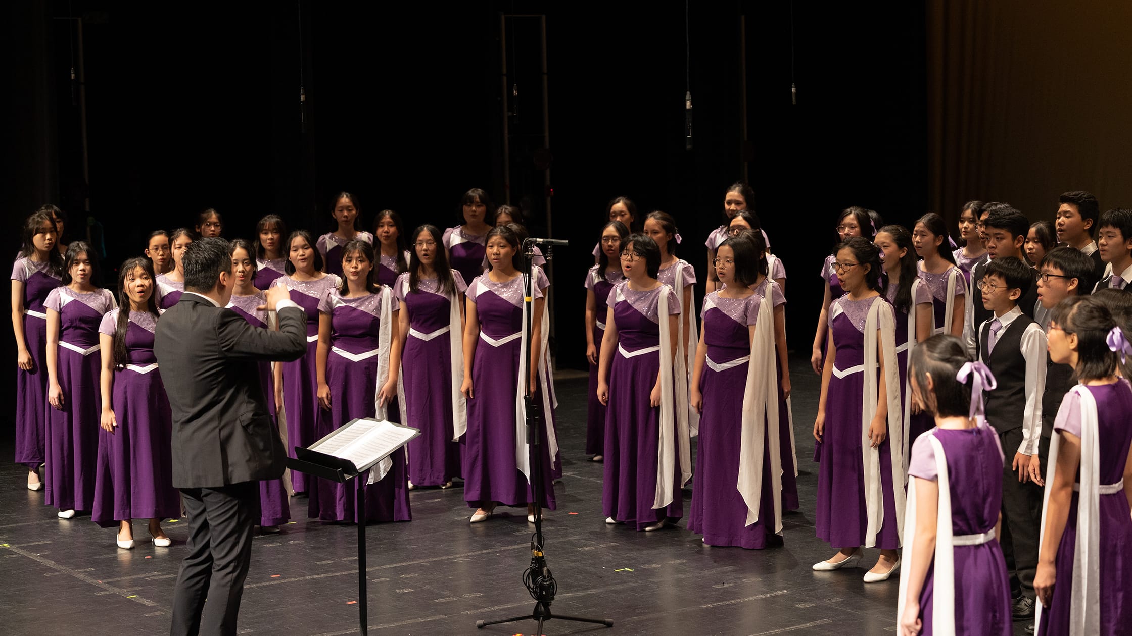 A conductor leads a youth choir in purple dresses with white scarves performing on a dark stage.