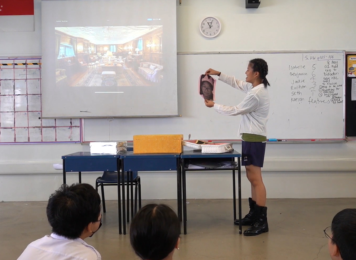 A student stands at the front of a classroom holding up a photo while a projection and whiteboard are visible behind.